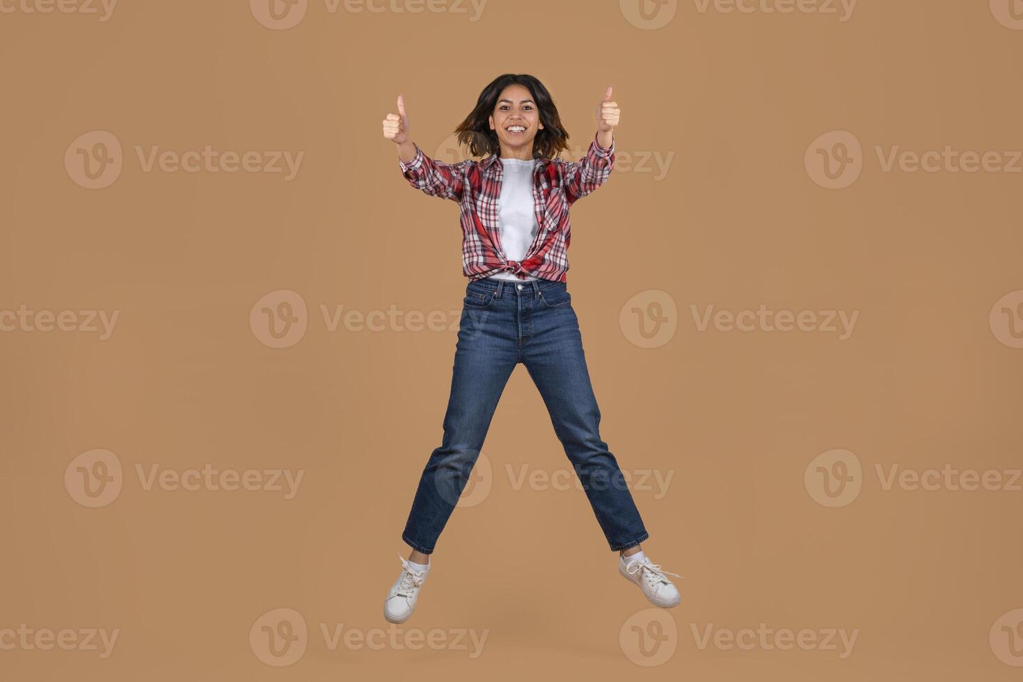 A young middle eastern woman with long, dark hair is jumping in the air with her arms outstretched and thumbs pointed upwards. She is wearing red and white plaid shirt, blue jeans, and white sneakers photo