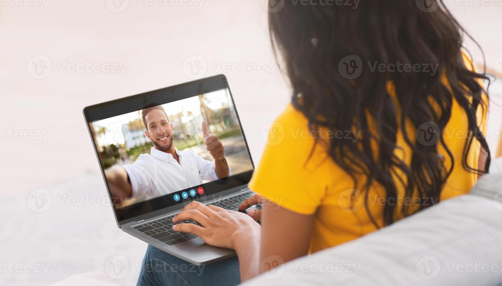 Black woman is seated on a couch, chatting with a man on her laptop. The man is giving a thumbs up in the call. She is looking at the screen and typing on the laptop keyboard. photo