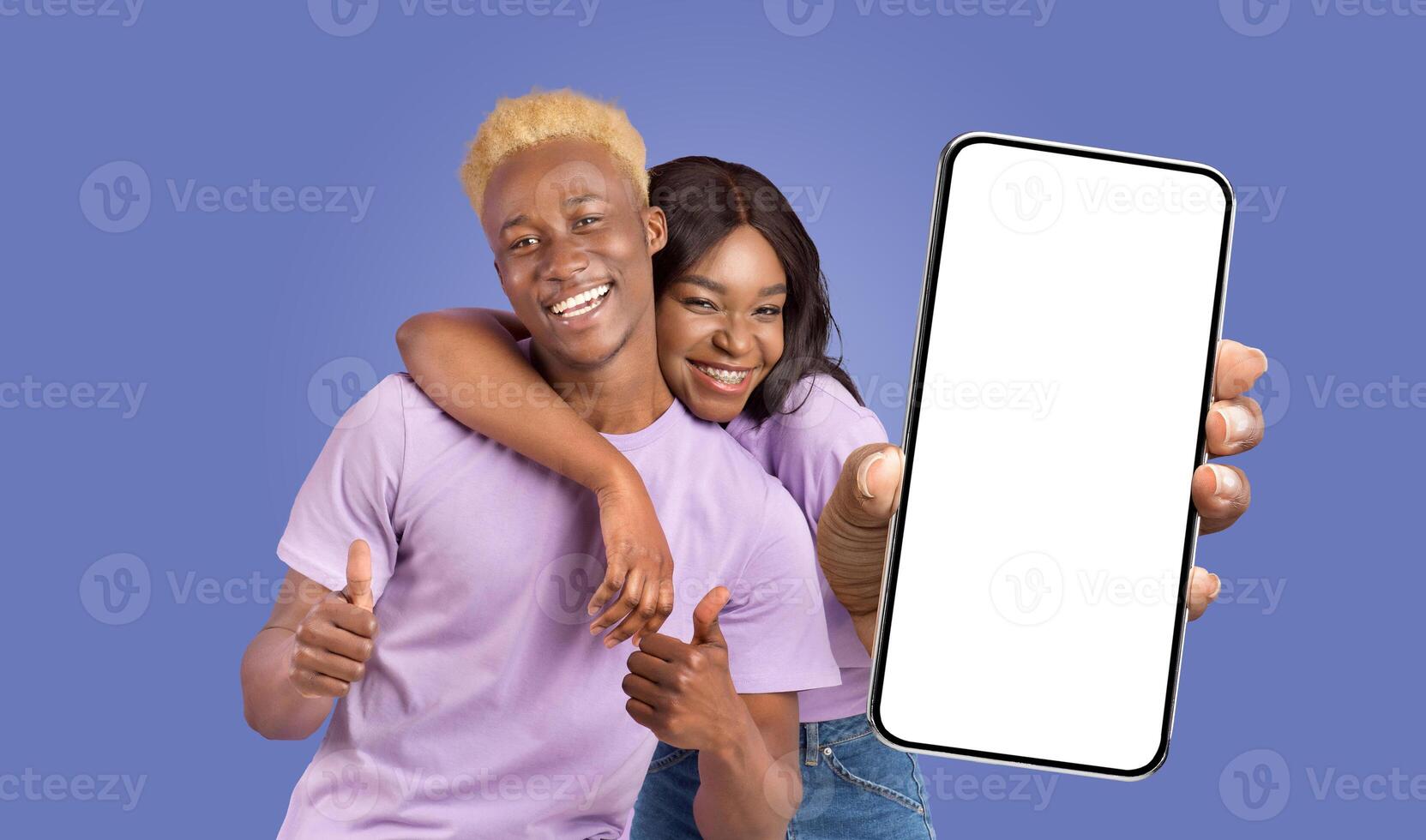 Black couple, both smiling, pose together in front of a blue background. The woman holds a smartphone with a white blank screen and points at it with her index finger. The man gives a thumbs up. photo