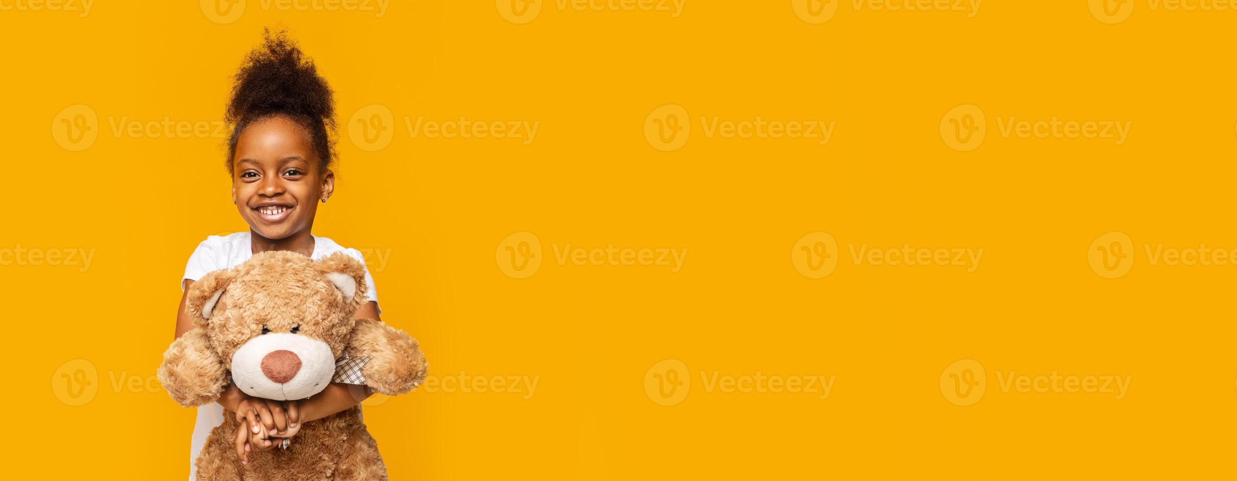 A joyful young African American girl stands confidently while hugging a large plush bear. She has a big smile on her face, radiating happiness. The background is vivid yellow, copy space photo