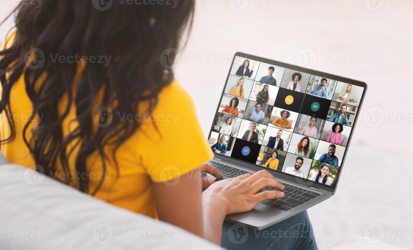 A young black woman is engaged in a virtual team meeting while working from her home office. She is seated, looking at a laptop screen displaying numerous colleagues via call photo