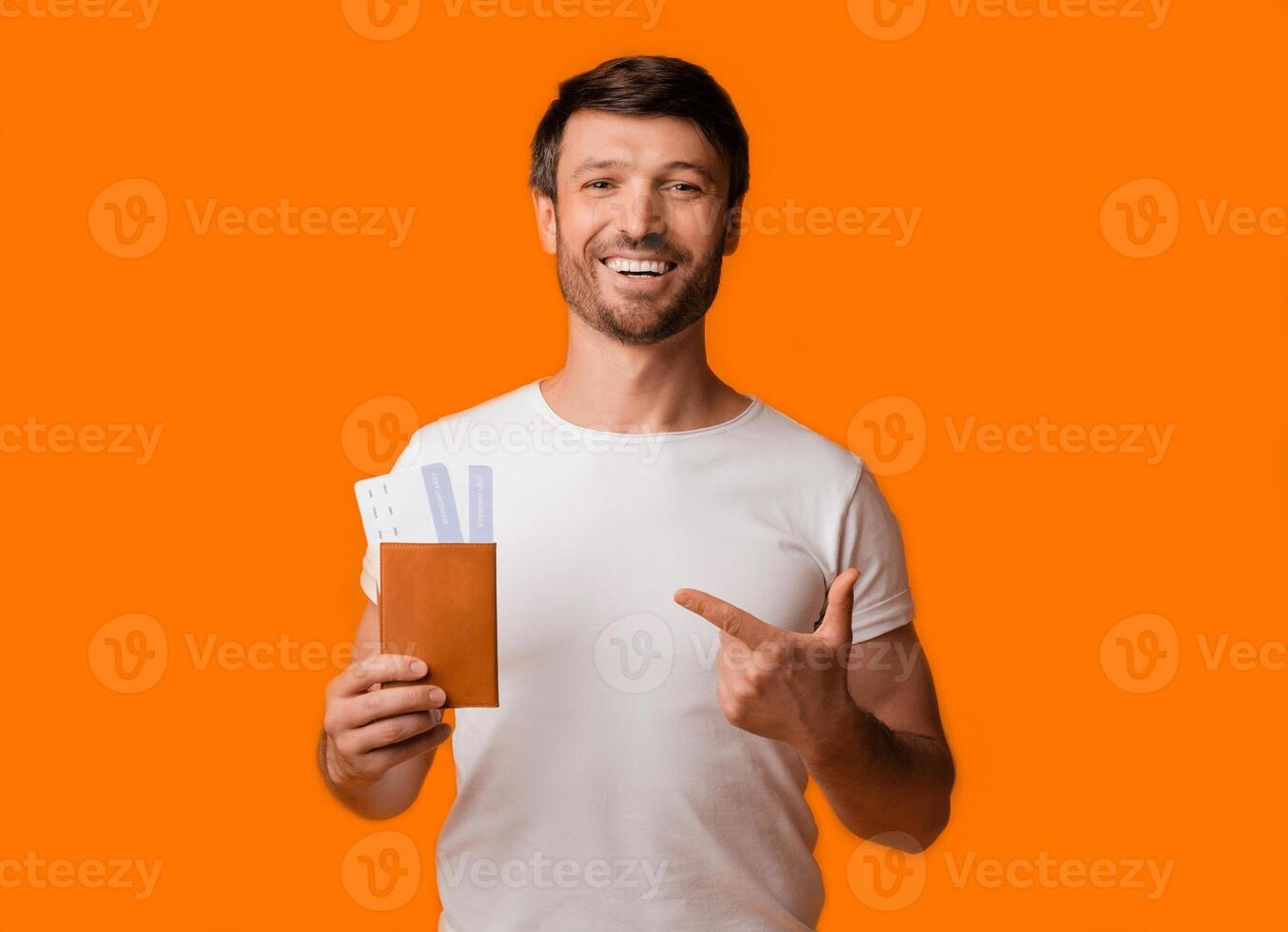 A smiling man in a white t-shirt holds a passport and airplane tickets in his left hand and points to the right with his right hand, against a bright orange background. photo