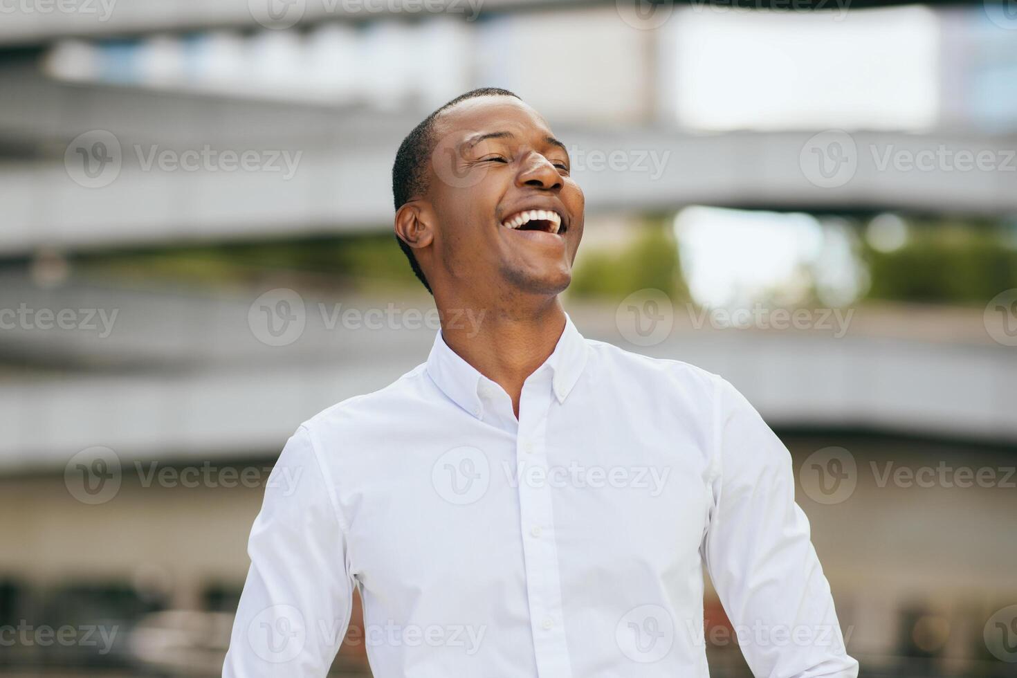 African American man wearing a white button-down shirt and laughing loudly with his head tilted back. He is standing in an urban setting with a blurred background of concrete and greenery photo