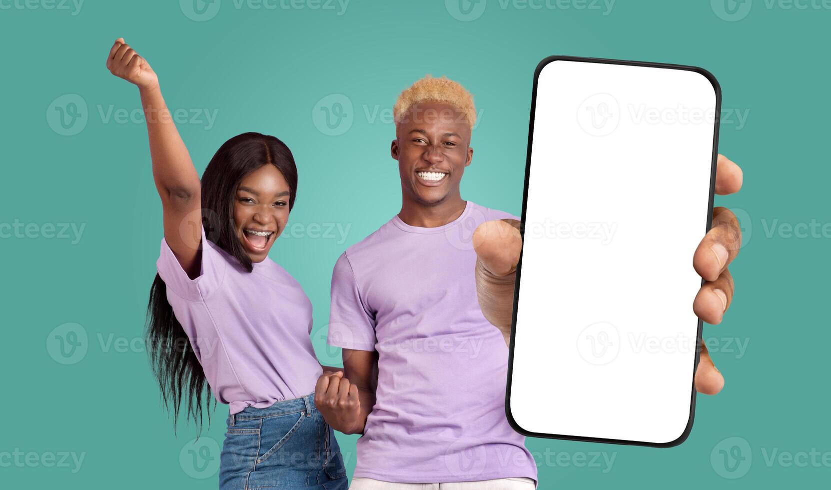 A young black couple in matching purple shirts stands in front of a teal background. The man holds a smartphone with a blank white screen towards the camera, while the woman celebrates success photo
