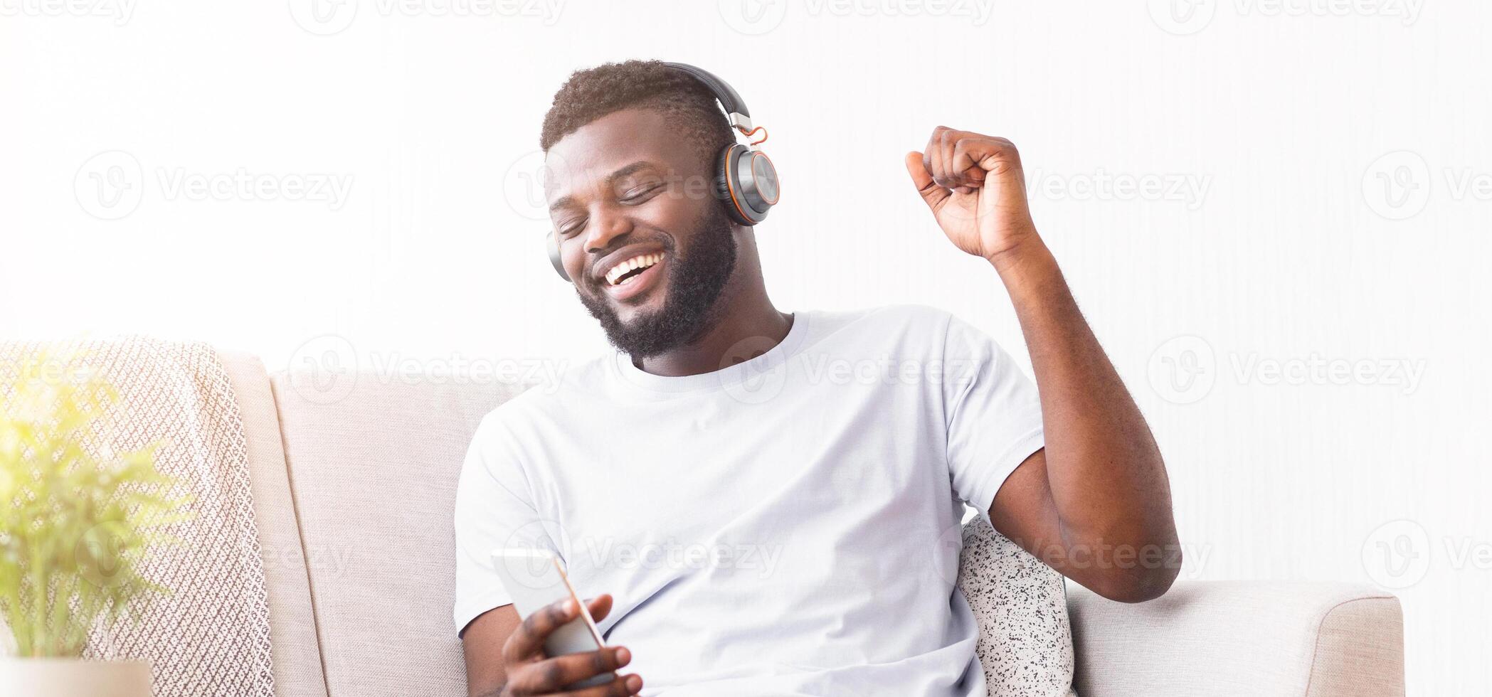 A happy Black man is sitting on a couch and listening to music on his smartphone through headphones. He is smiling and bobbing his head to the music. photo