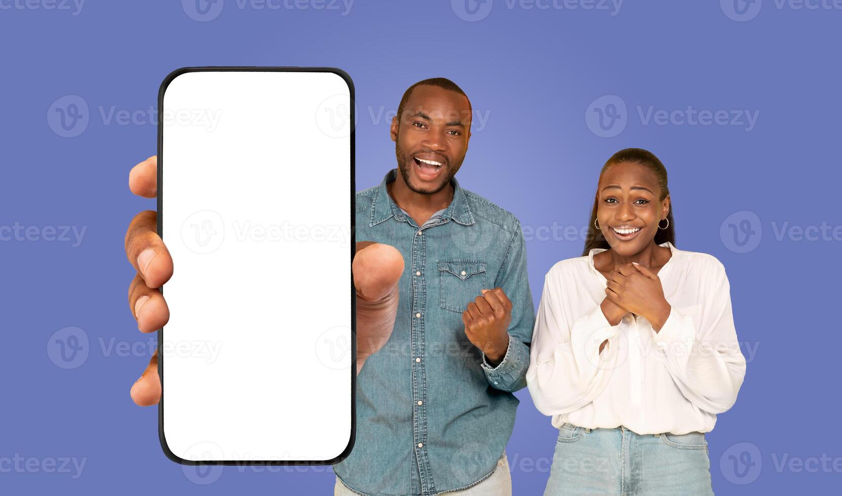 Black man and woman are standing in front of a blank smartphone screen held by an unseen hand. They are both smiling excitedly photo