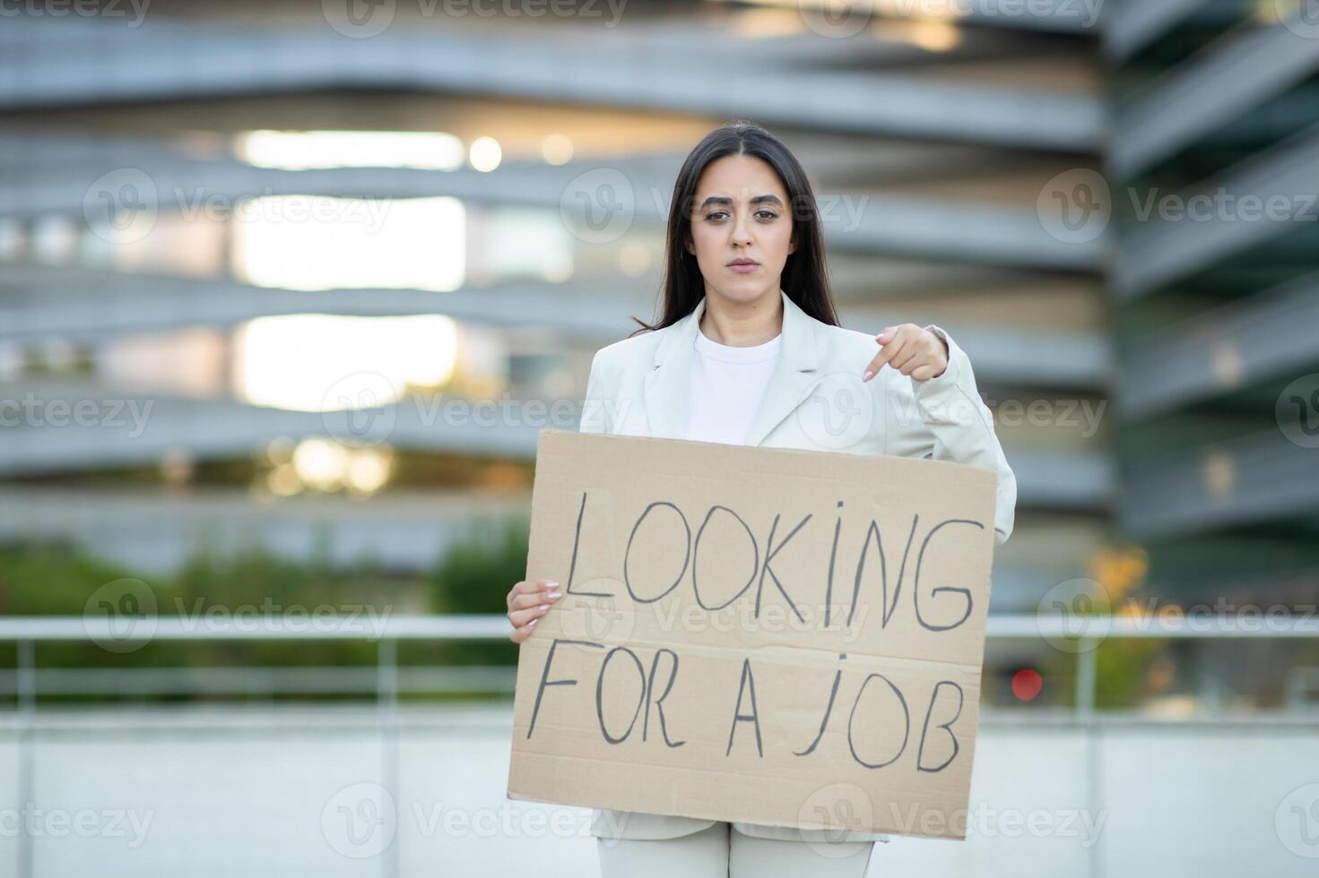 A woman wearing a white blazer stands in front of a modern city building, holding a cardboard sign that reads Looking For A Job. She points at the sign with her right hand photo