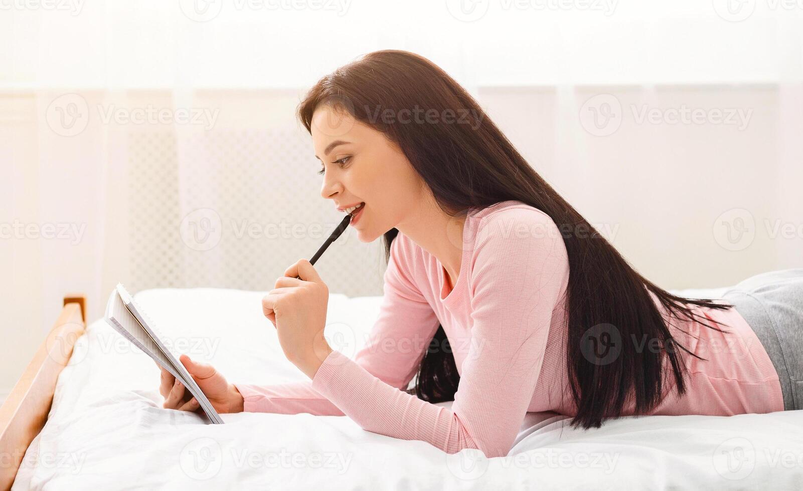 A young woman with long, dark hair is lying on bed in bright room, intently writing in notebook with pen. Her expression shows focus and contemplation as she appears to be planning or journaling. photo