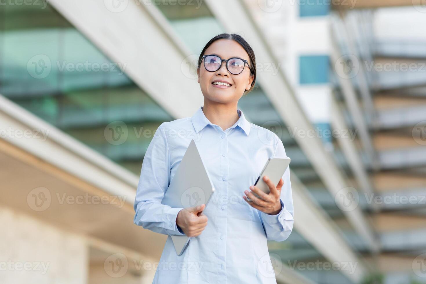 A young Asian woman confidently walks outside a contemporary building while holding a tablet in one hand and a smartphone in the other. She wears glasses and a light blue button-up shirt photo