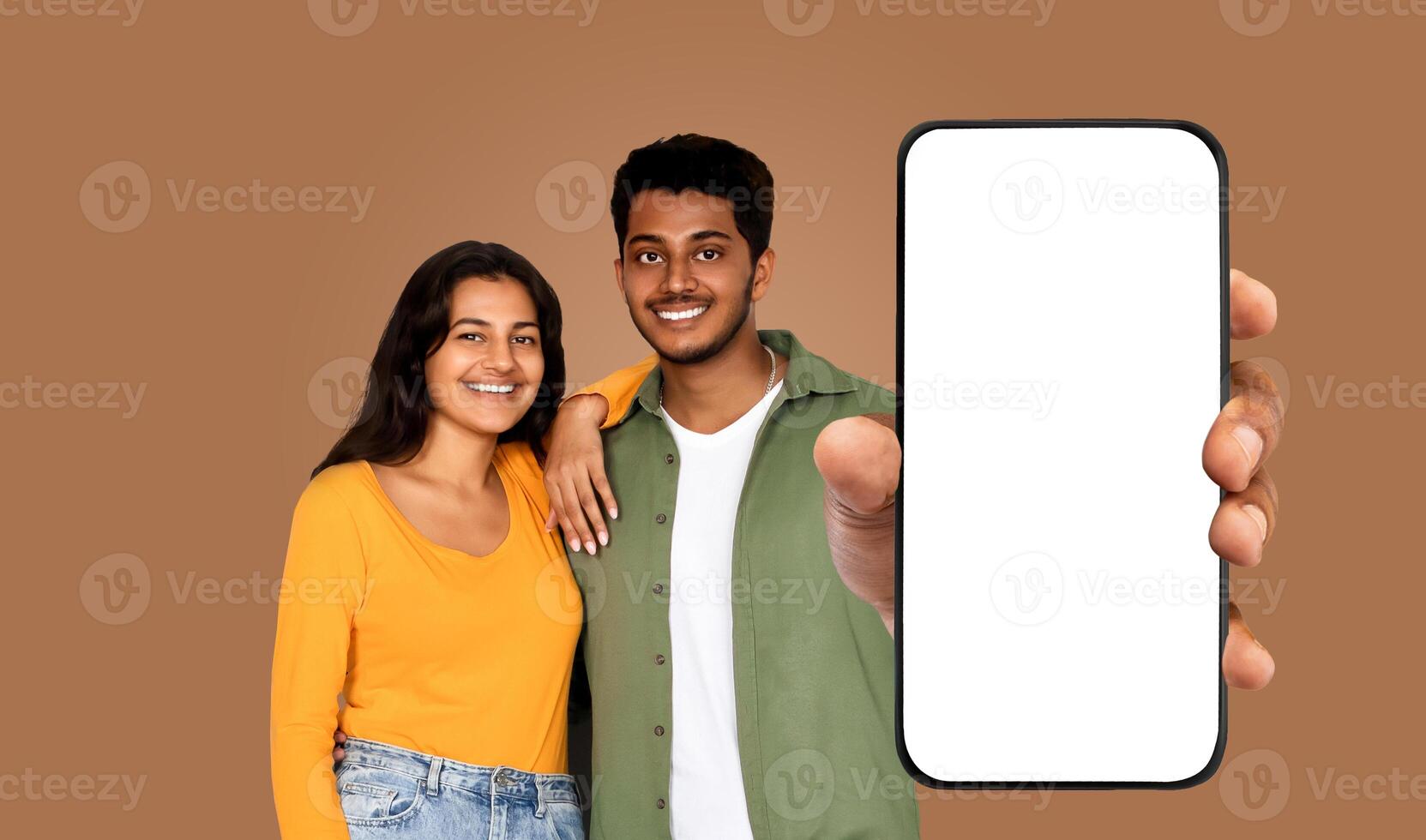A young Indian couple stand against a brown background. The man is holding a smartphone with a blank screen, pointing at the screen with his index finger photo