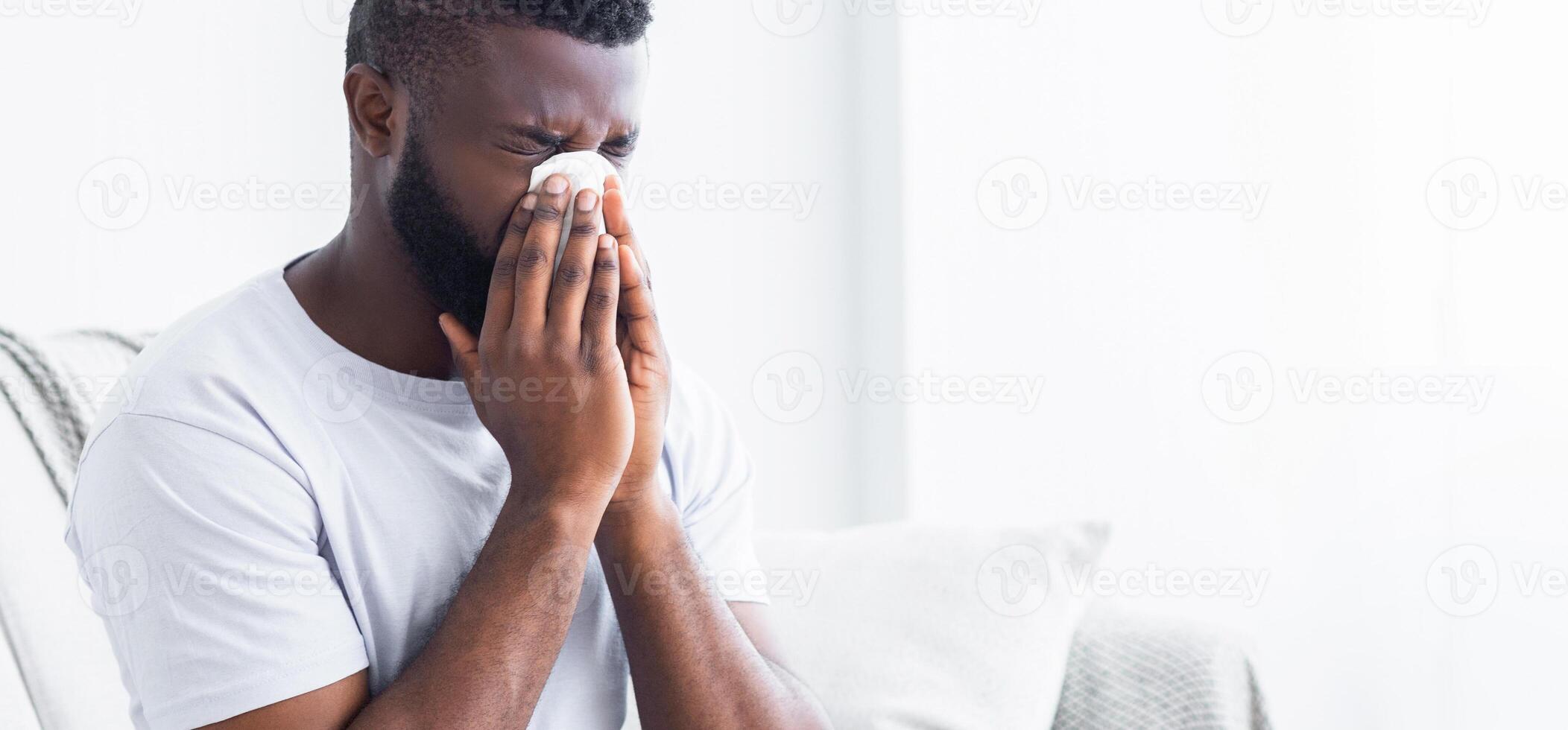 African American man sits on a bed, his head bowed, blowing his nose into a tissue. He appears to be suffering from a cold or allergies. The room is well-lit and has a white background, copy space photo