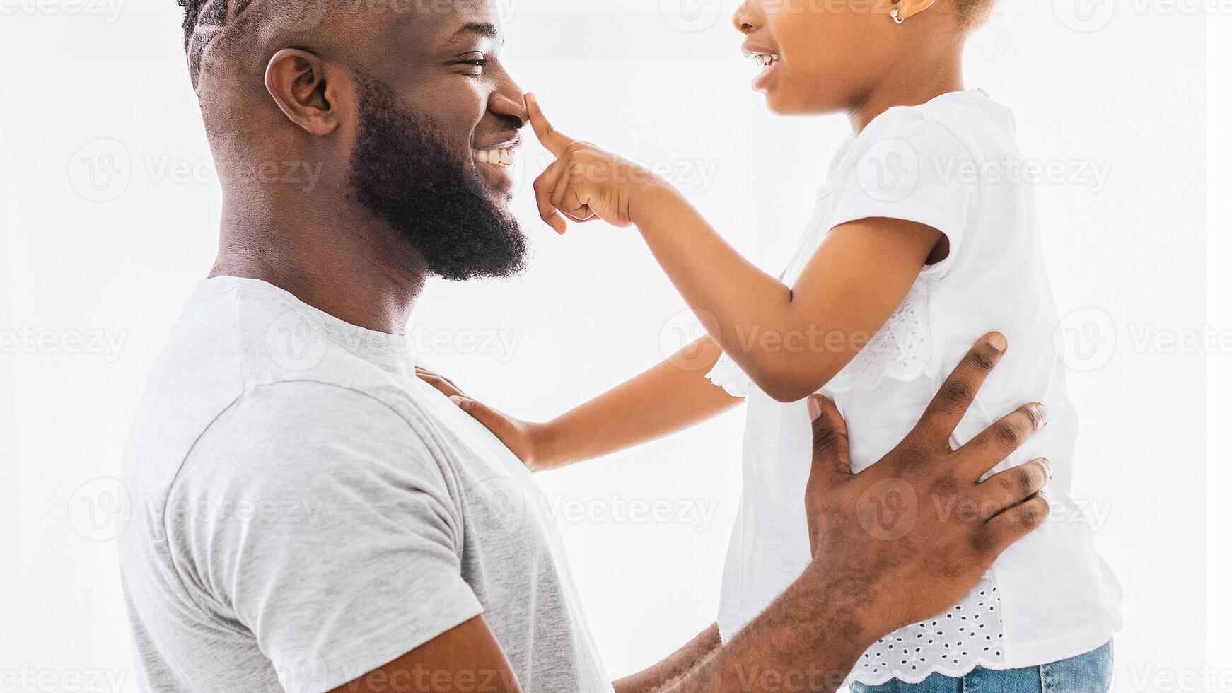A happy Black father and daughter are playing together indoors. The daughter is touching her fathers nose while he smiles and looks at her. photo