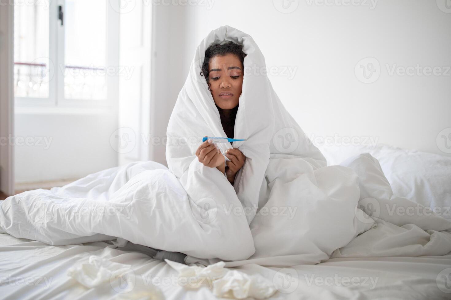 A young Hispanic woman appears to be unwell, sitting in her bed wrapped in a white blanket with tissues scattered around her. She is holding a thermometer, presumably checking her temperature photo