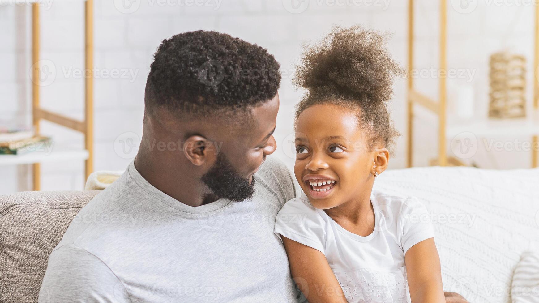A close-up image of a happy Black father and daughter laughing together on a couch in a living room. The two are enjoying each others company and are clearly having a good time. photo