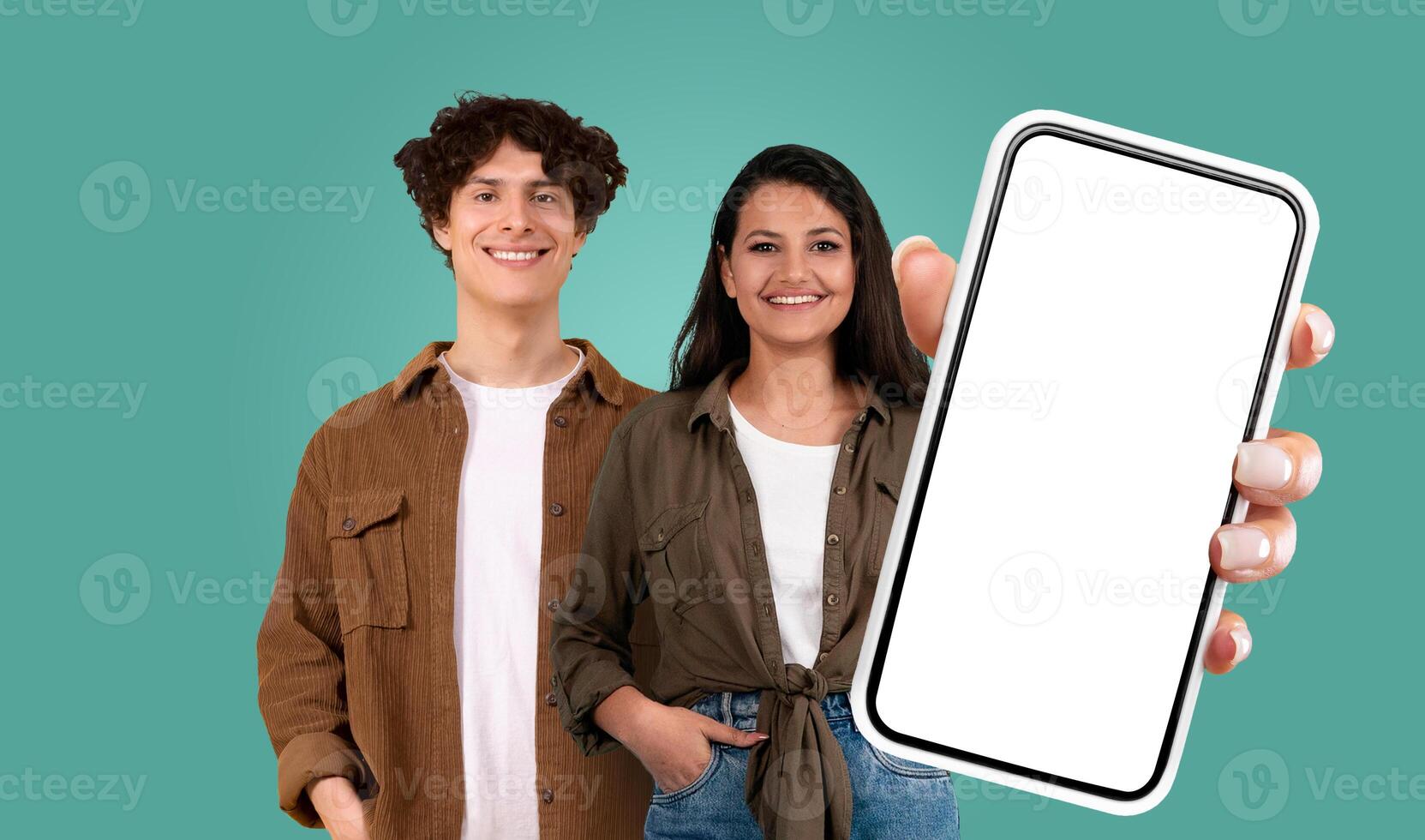 Multiracial couple stand against a teal backdrop. The woman holds a large smartphone with a blank screen in her right hand, extending it out towards the viewer. Both are smiling. photo