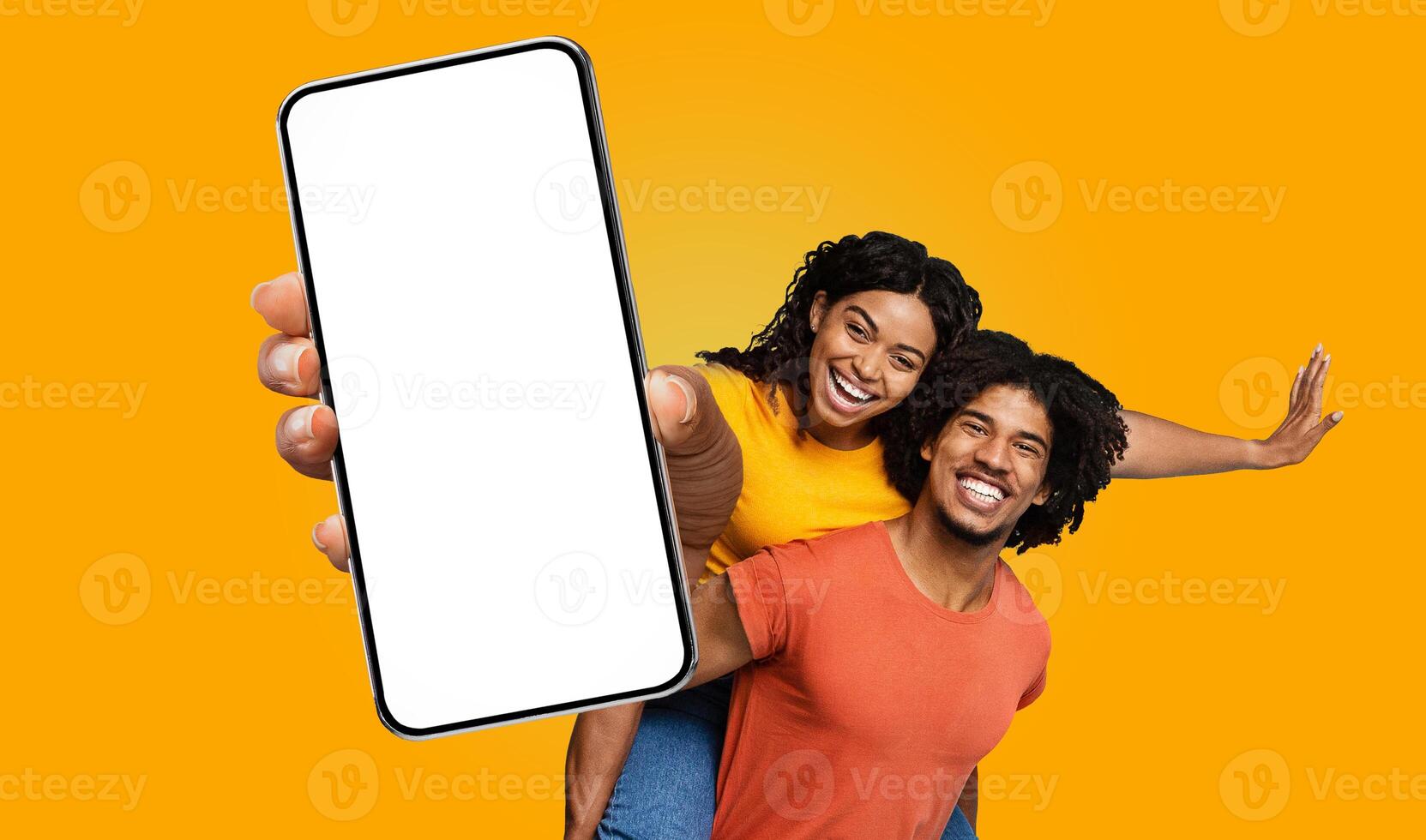 A young black woman in a yellow shirt is giving a piggyback ride to a man in a red shirt, both smiling broadly. The woman is pointing at the blank screen of a large smartphone photo