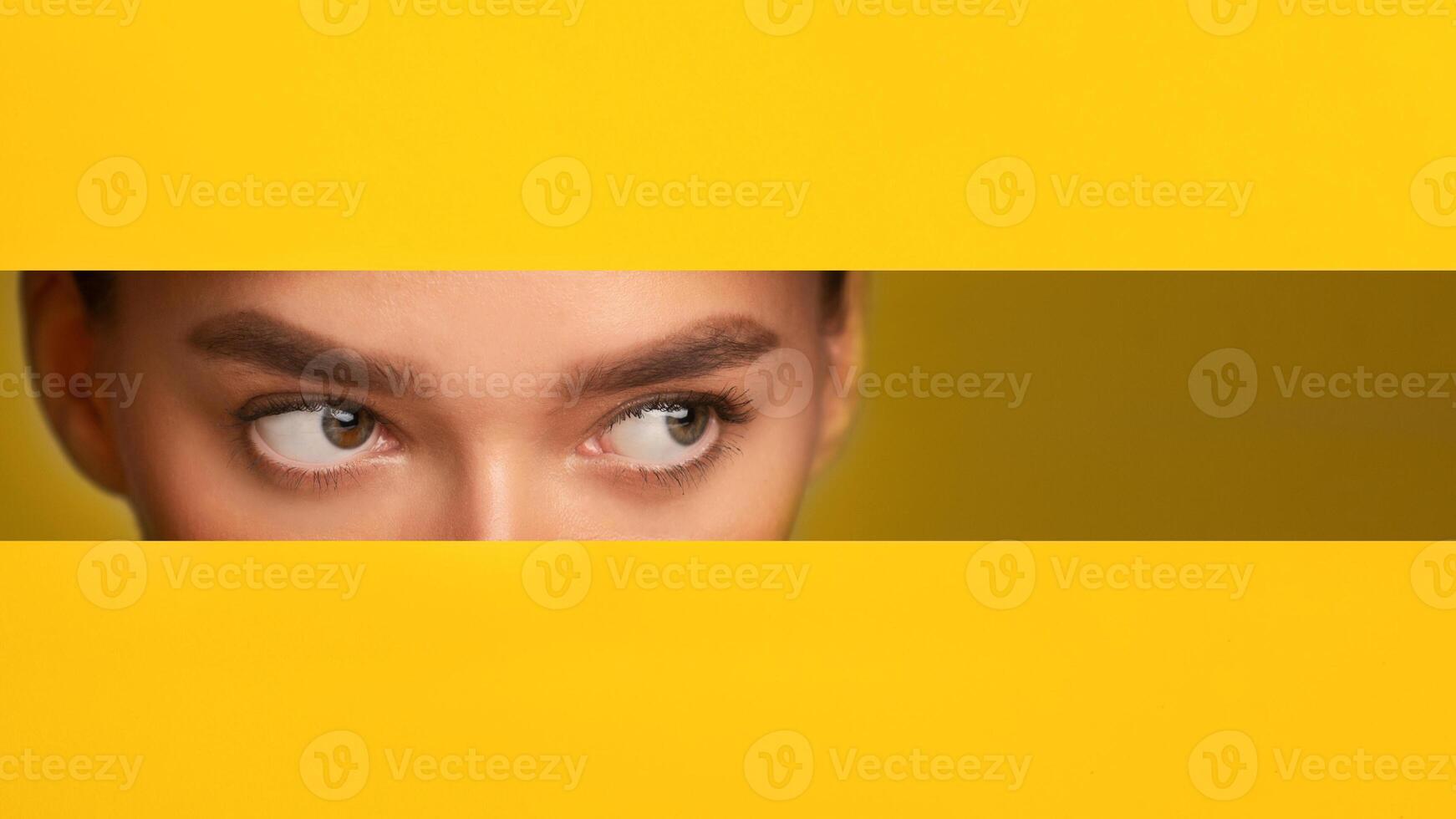 A close-up of a woman eyes filling the frame, set against a vibrant yellow paper wall background. The woman features are visible, showcasing her eyes, nose, copy space photo