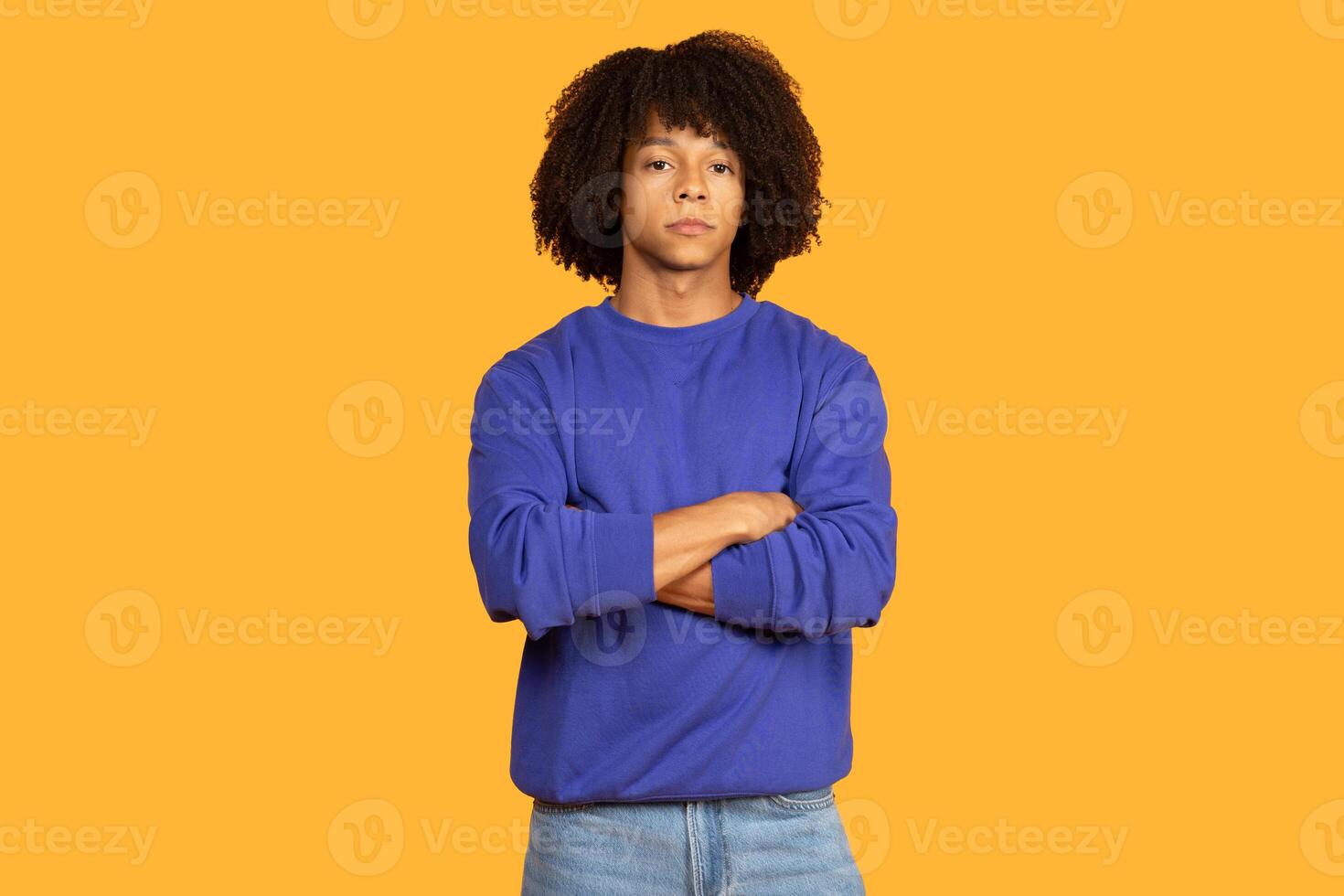 A young man stands confidently with arms crossed, showcasing a relaxed demeanor. The vibrant yellow background enhances the subject's presence, drawing attention to his expression and outfit. photo