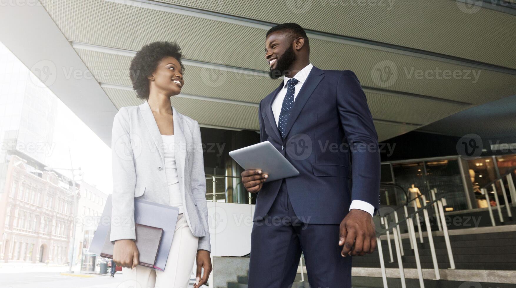 Two smiling african american colleagues discussing new project while going out of office building, copy space photo