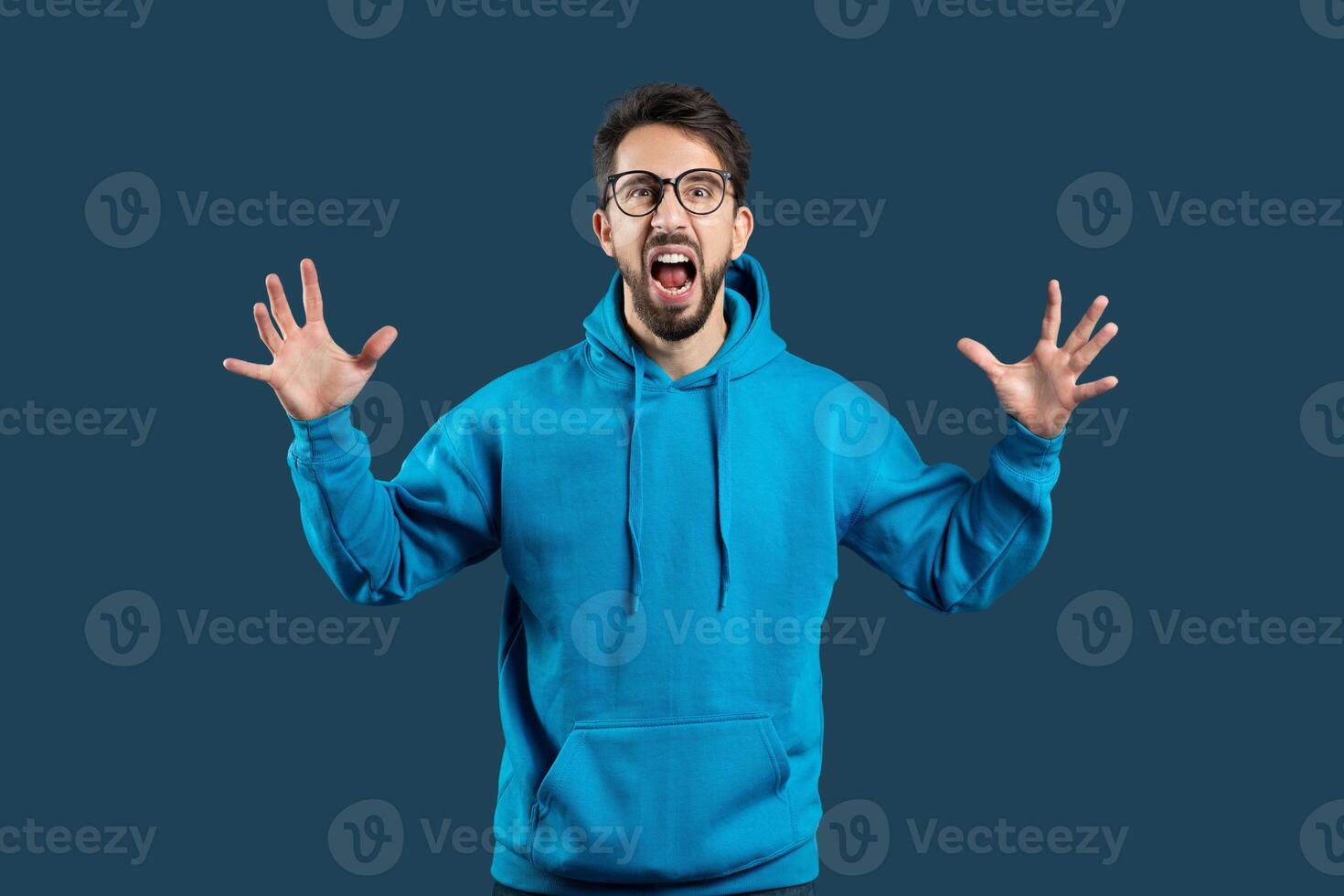 A delighted young man with glasses is standing against a dark blue backdrop, his mouth wide open as if shouting with joy or surprise. He is wearing a bright blue hoodie and has raised his hands photo