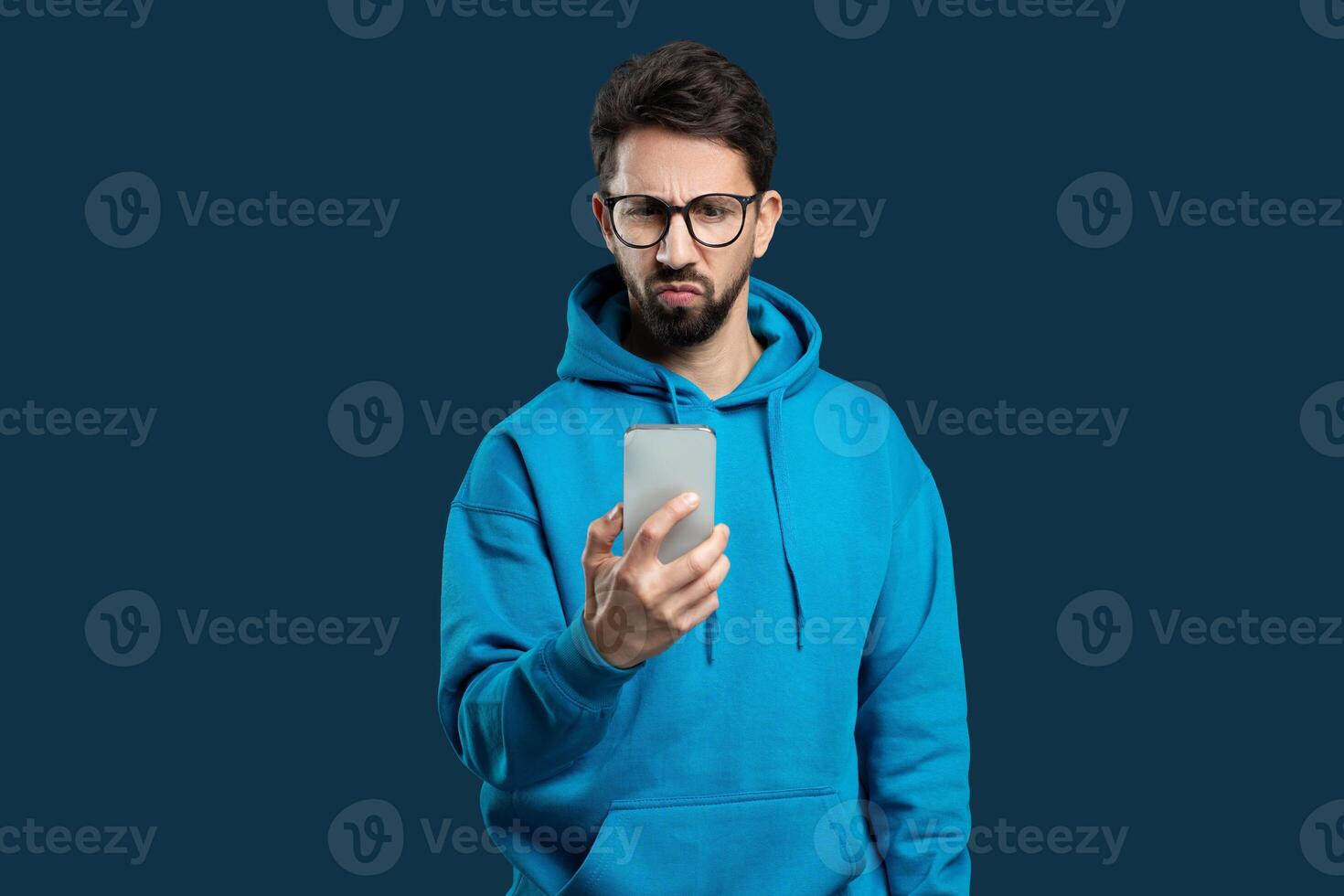 A young, bearded man wearing glasses and a blue hoodie stands against a solid blue background, looking at his smartphone with a skeptical and slightly confused expression. photo