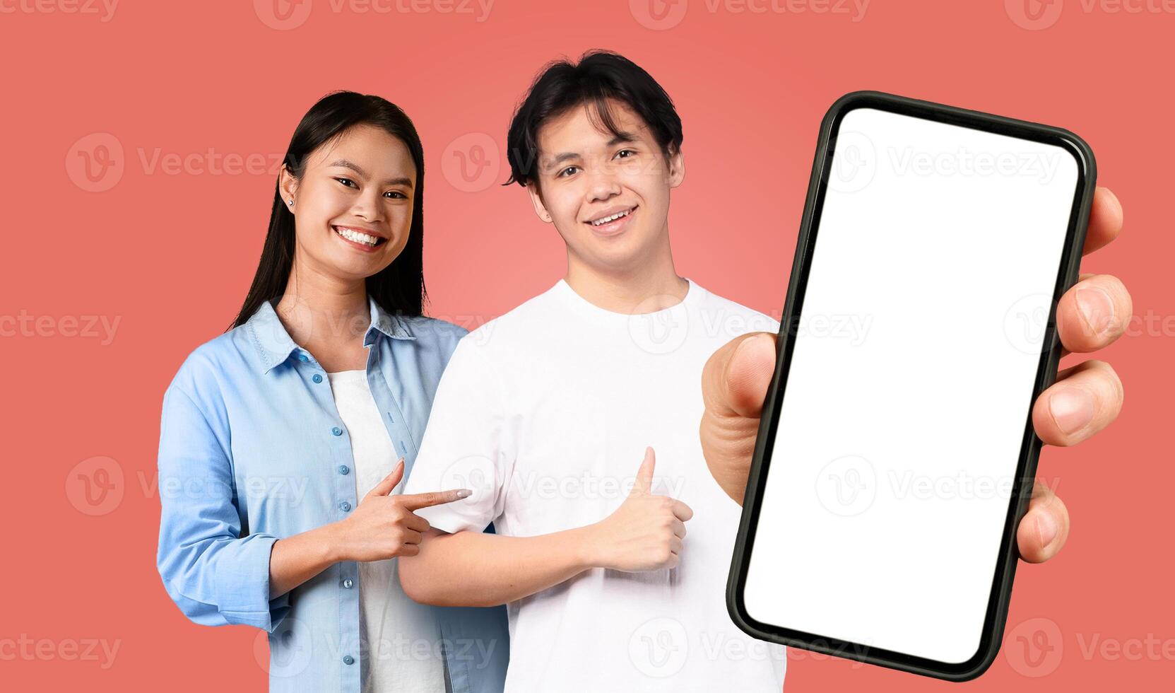 Two young Asian people stand in front of a red background. The man is holding a smartphone with a blank screen in his right hand, and he is pointing at the screen with his left hand. photo