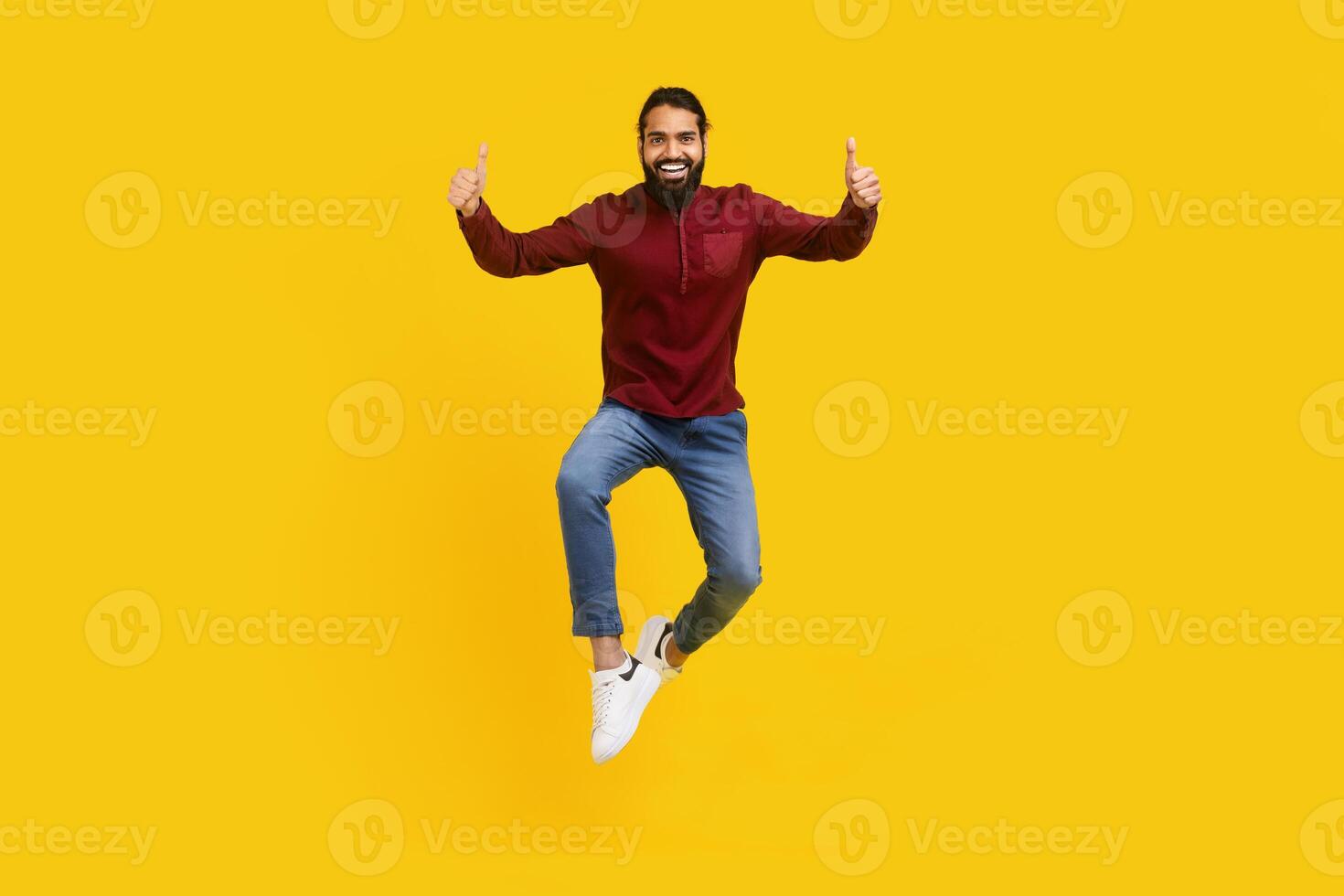 Indian man is captured mid-air, jumping energetically with his hands raised in thumb ups gesture, yellow studio background photo