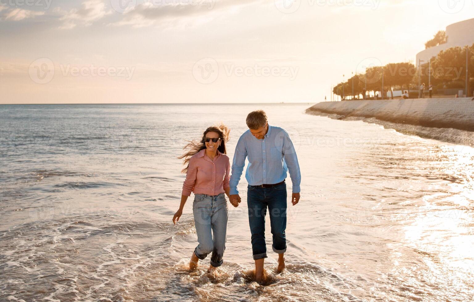 A couple strolls along the shoreline, holding hands as light waves wash over their feet. The sun sets in the background, casting a warm glow around them, enhancing the romantic atmosphere. photo