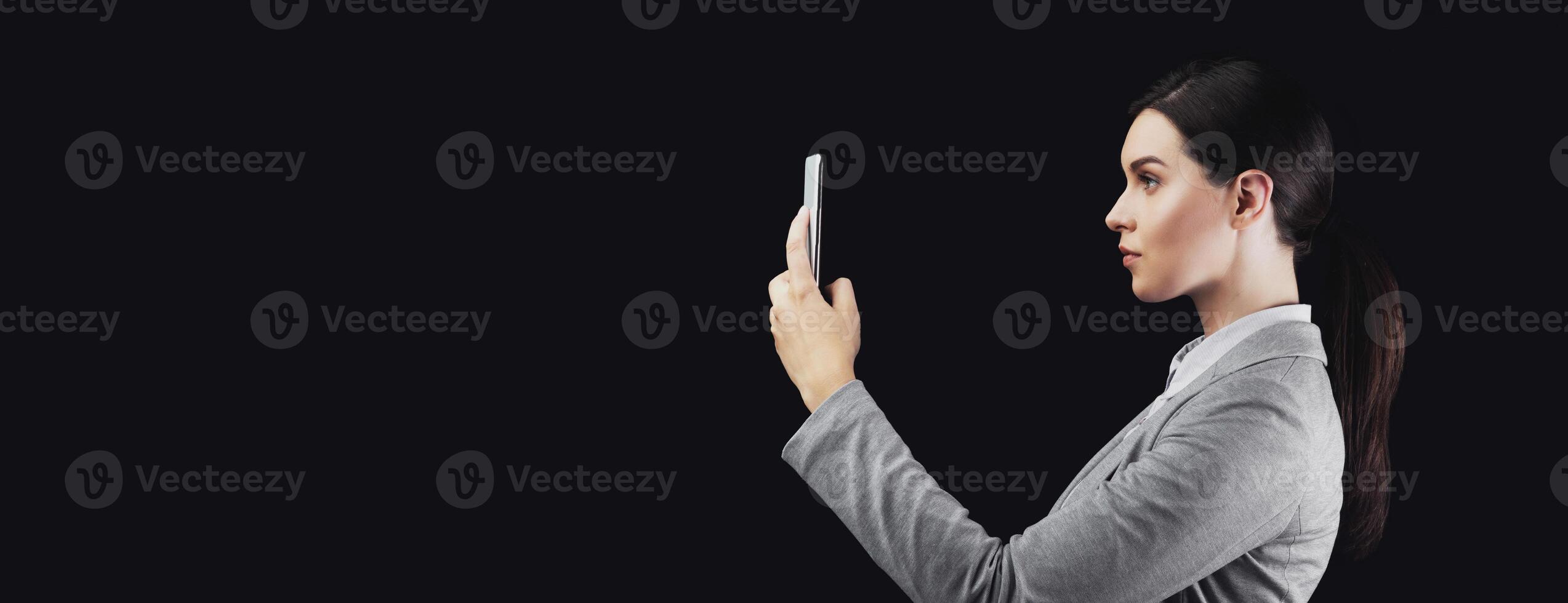 A woman in a grey blazer holds a smartphone in front of her face, looking directly at it. The background is black. photo