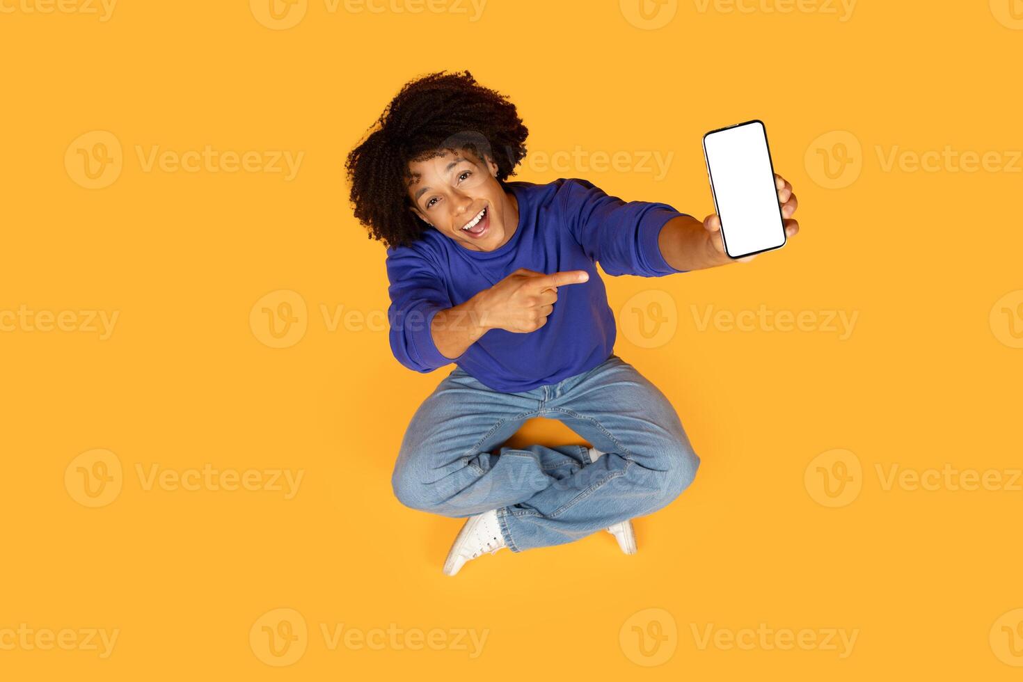 A guy is seated on the floor, his finger extended to point at a cell phone with white screen in his hand. He seems to be engaged with the device, possibly showing something on the screen. photo