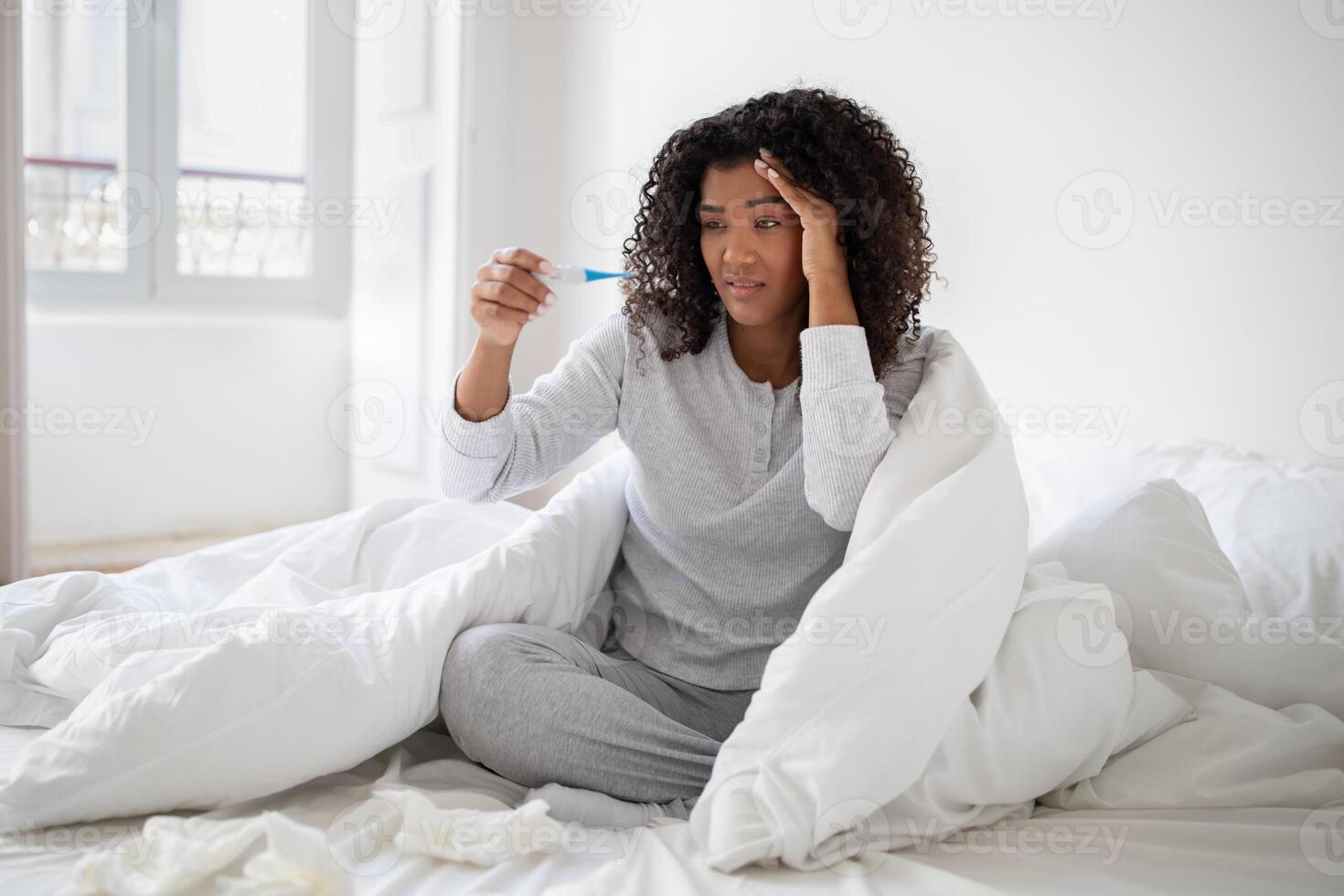 A young Hispanic woman wrapped in a white blanket is sitting up in bed with a concerned expression as she looks at a thermometer. photo
