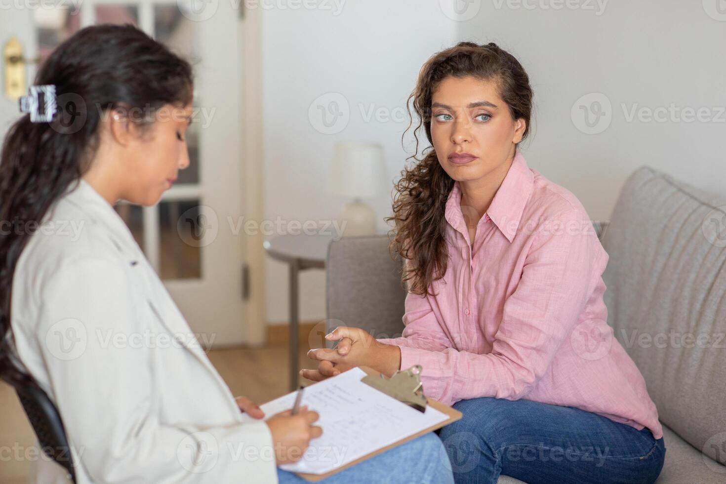 Unhappy young woman sits on a couch, listening intently to a therapist, who takes notes on a clipboard. photo
