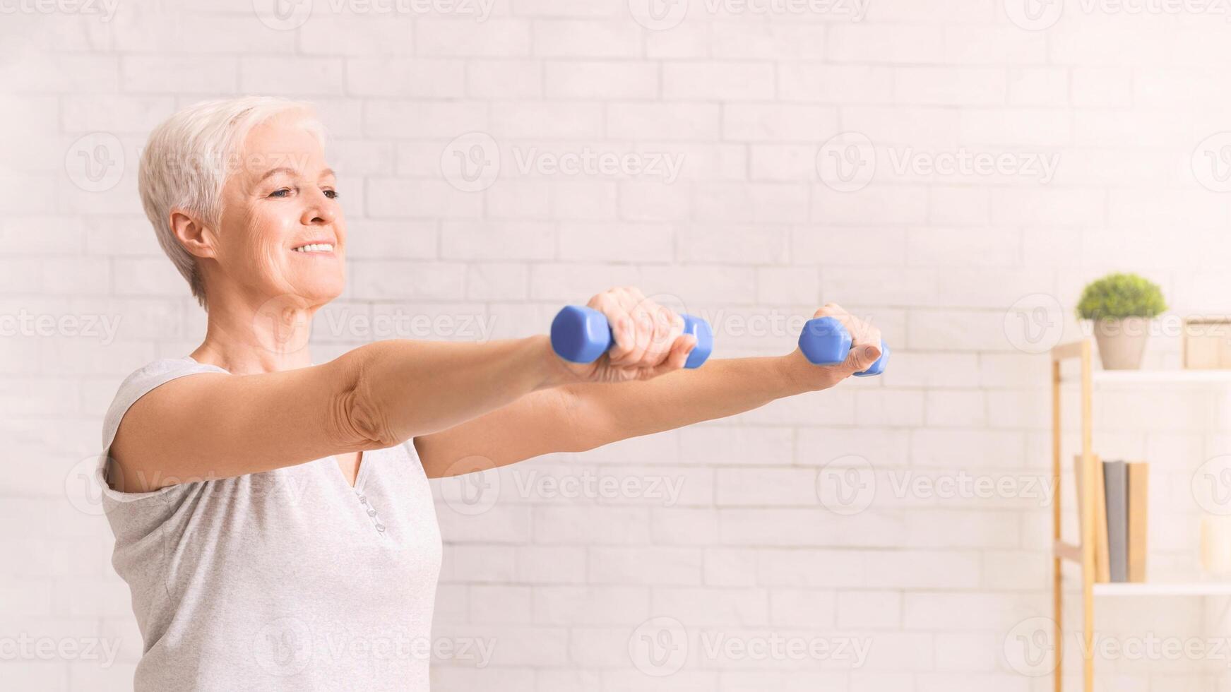 An older woman is actively exercising in a white room, lifting dumbbells to strengthen her muscles and improve her physical fitness photo