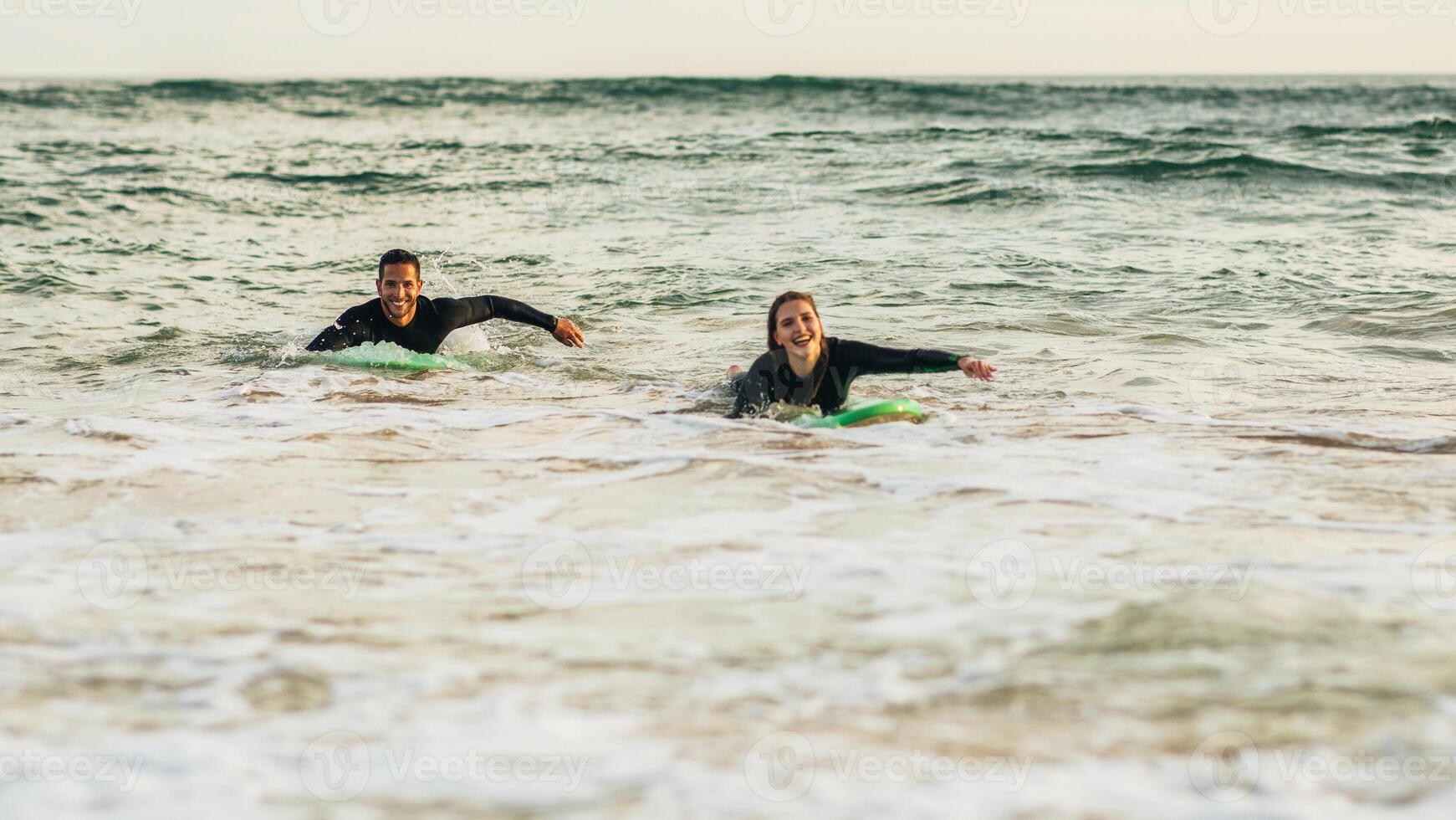 A couple in black wetsuits paddle board on calm ocean waves. The sun is setting in the background, casting a warm glow on the water. They are smiling and appear to be enjoying their time together. photo