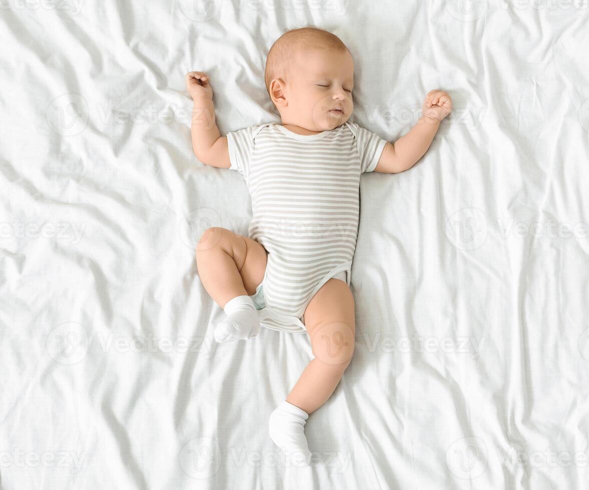 Adorable newborn baby in bodysuit napping on white blanket with his arms up, top view, copy space photo