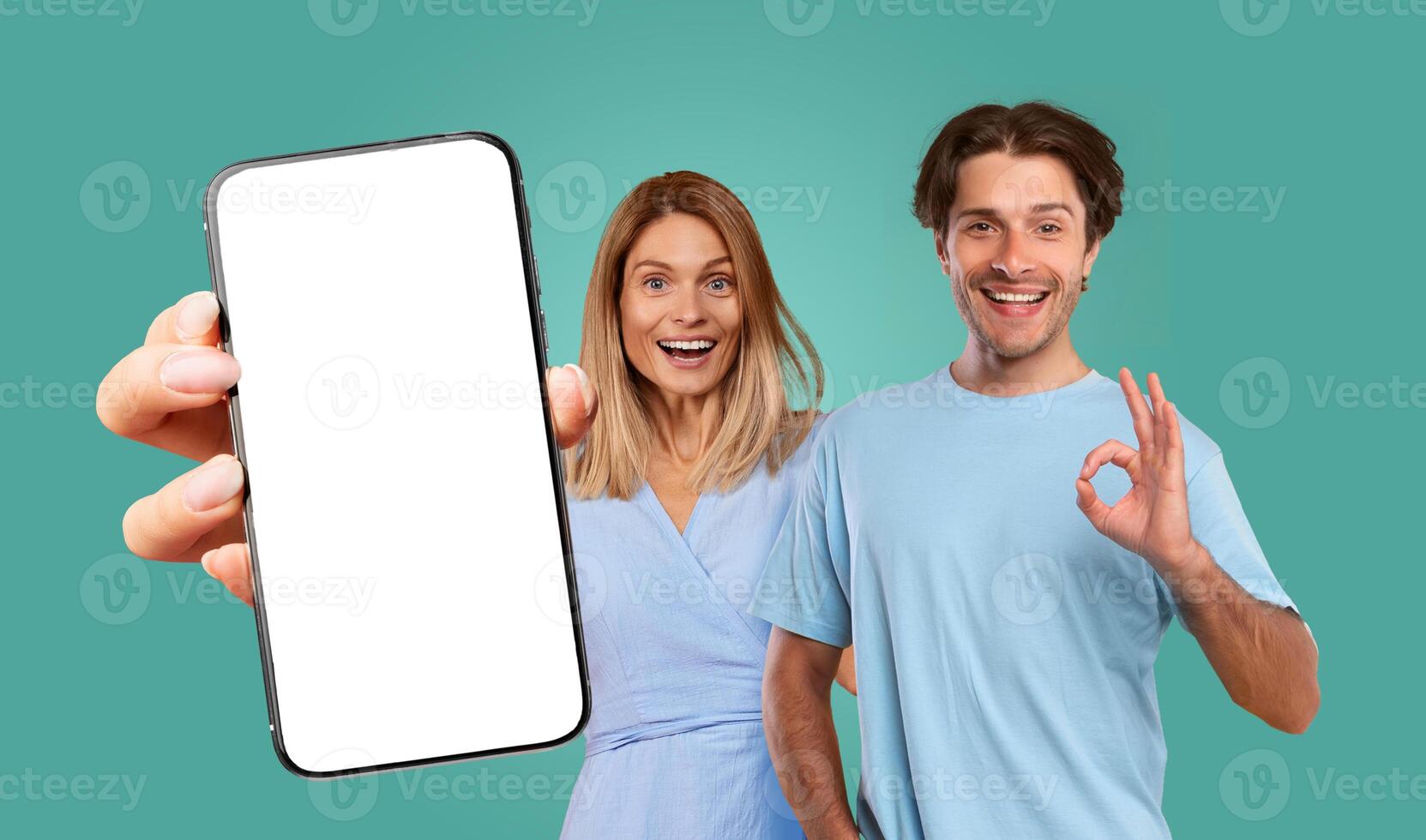 A couple poses happily in front of a teal background. The woman holds a smartphone with a blank screen and is smiling excitedly, while the man gives a thumbs up, showing his approval. photo
