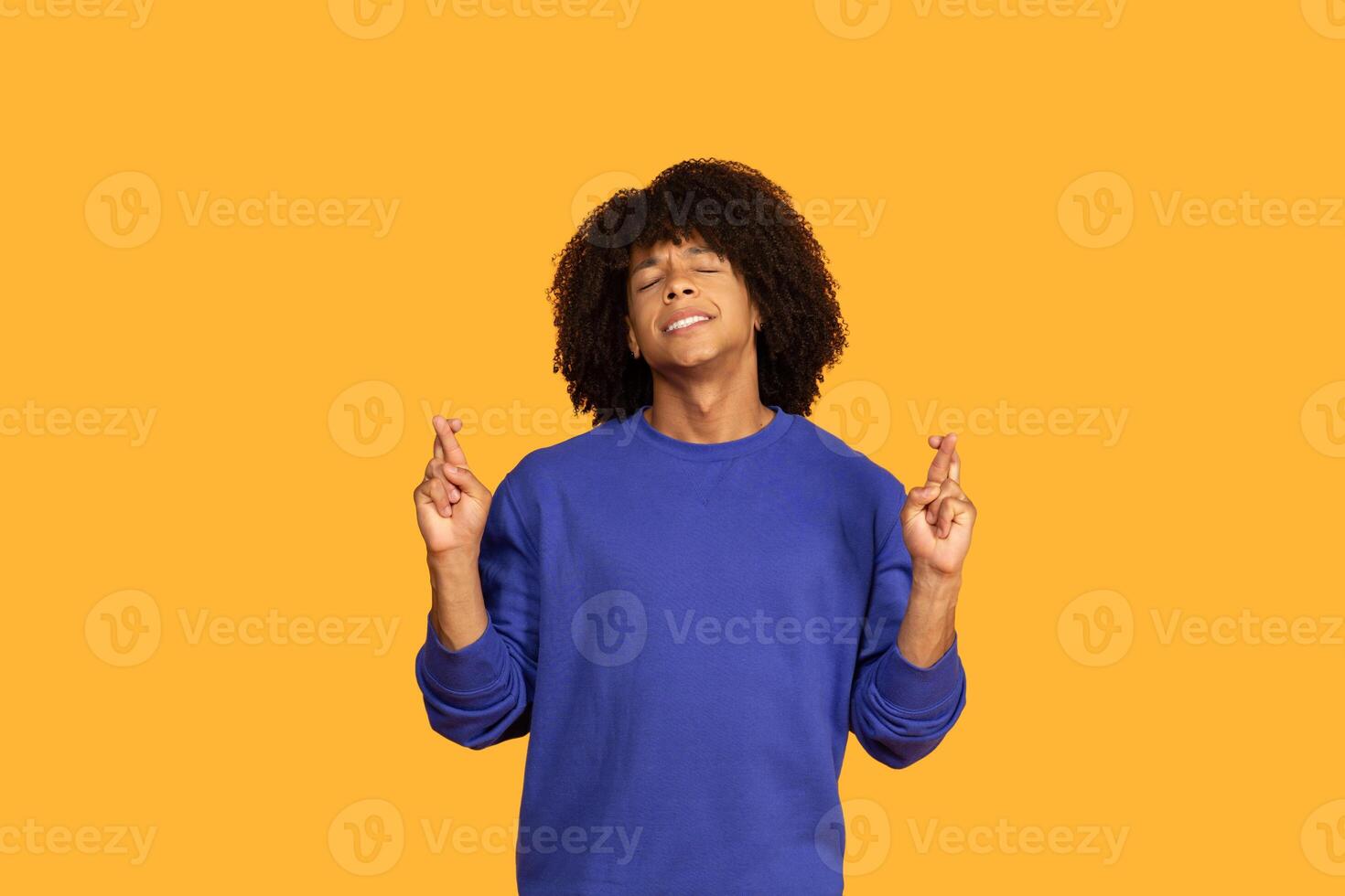 A young man stands against a vibrant yellow background, smiling cheerfully with his eyes closed. He has curly hair and is crossing his fingers, conveying a sense of hope and excitement. photo