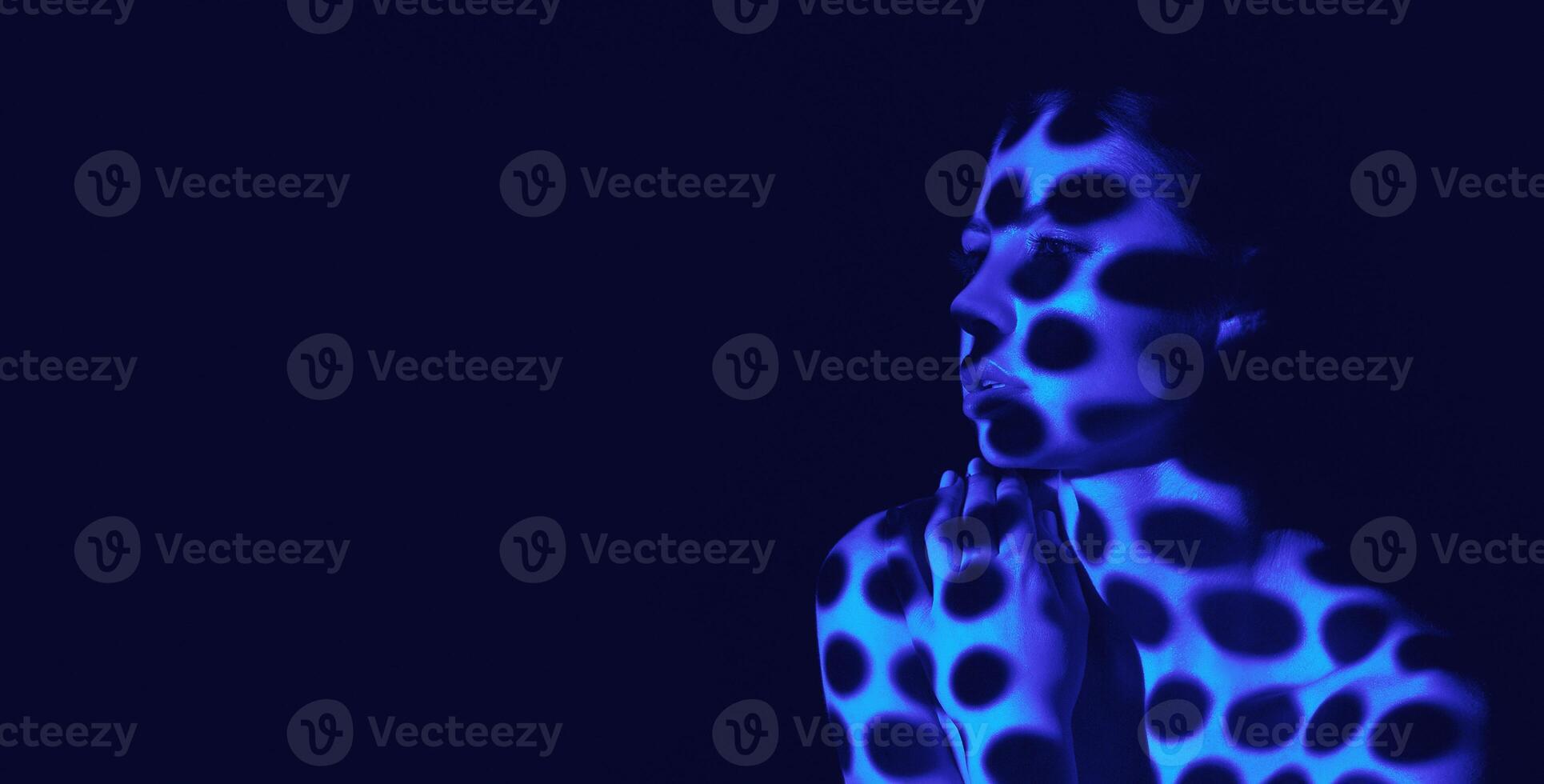 The image shows a womans face and upper body illuminated by blue light. The light creates a pattern of dots on her skin, like a pointillist painting. Her hands clasped in front of her, copy space photo