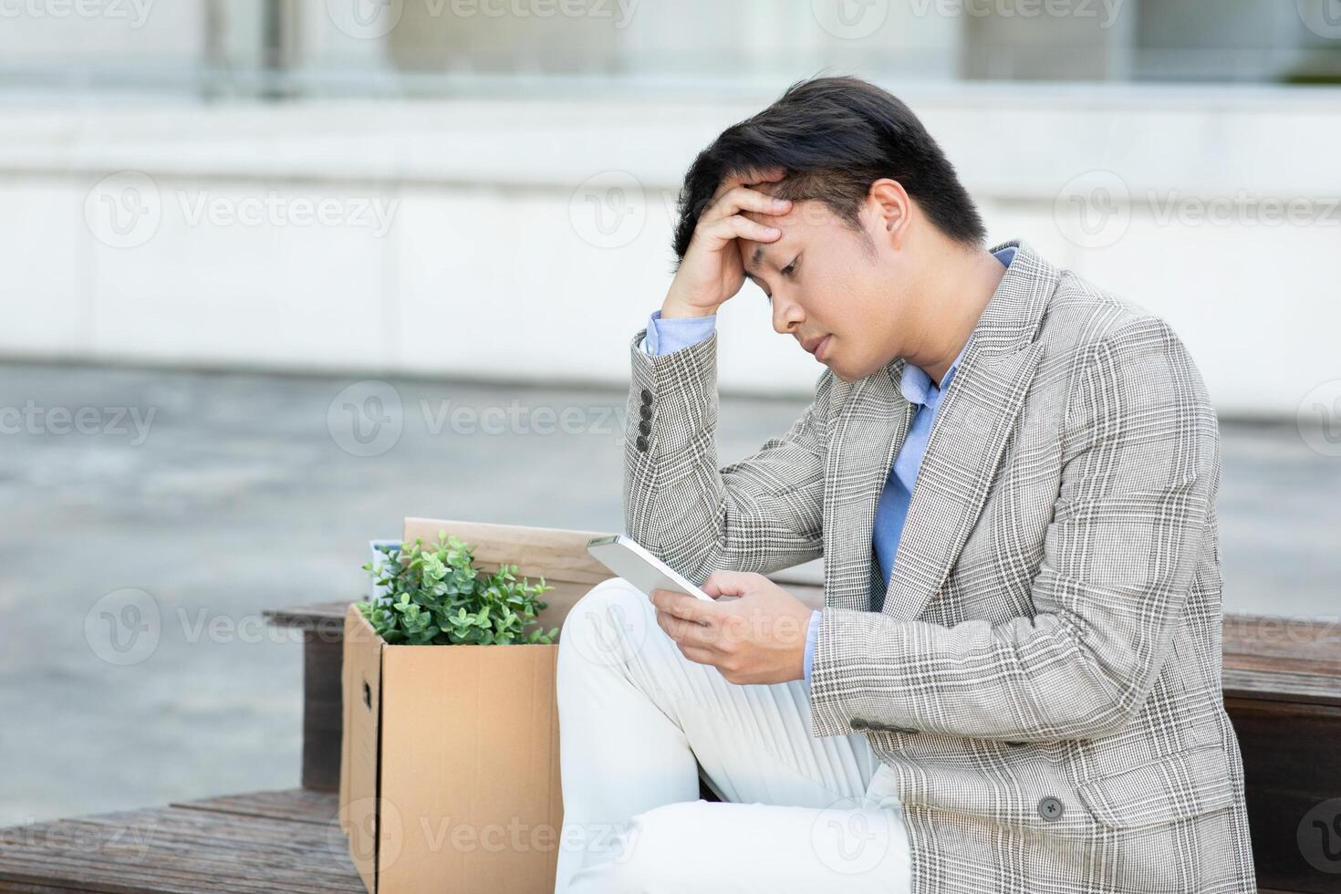 Asian young man in a grey plaid blazer sits on a bench outside with a cardboard box beside him. He is holding a phone in his hand and has a worried expression on his face, copy space photo