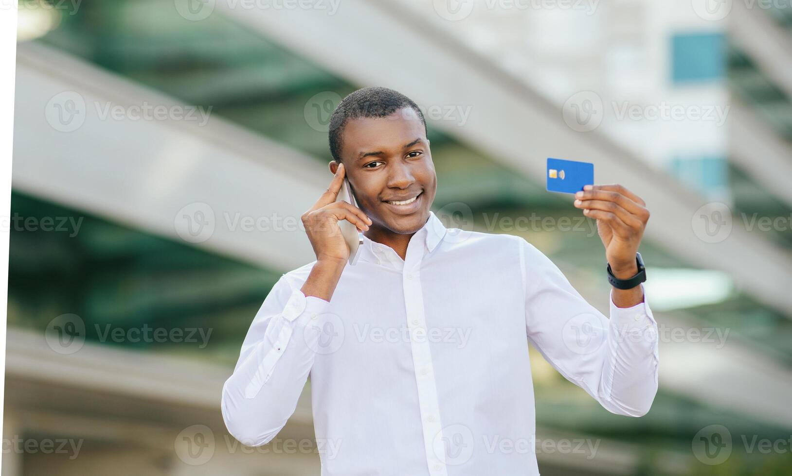 Black man in white shirt stands outside and smiles while holding a credit card in his right hand and talking on his cell phone. He is looking at the camera and standing in front of a modern building. photo