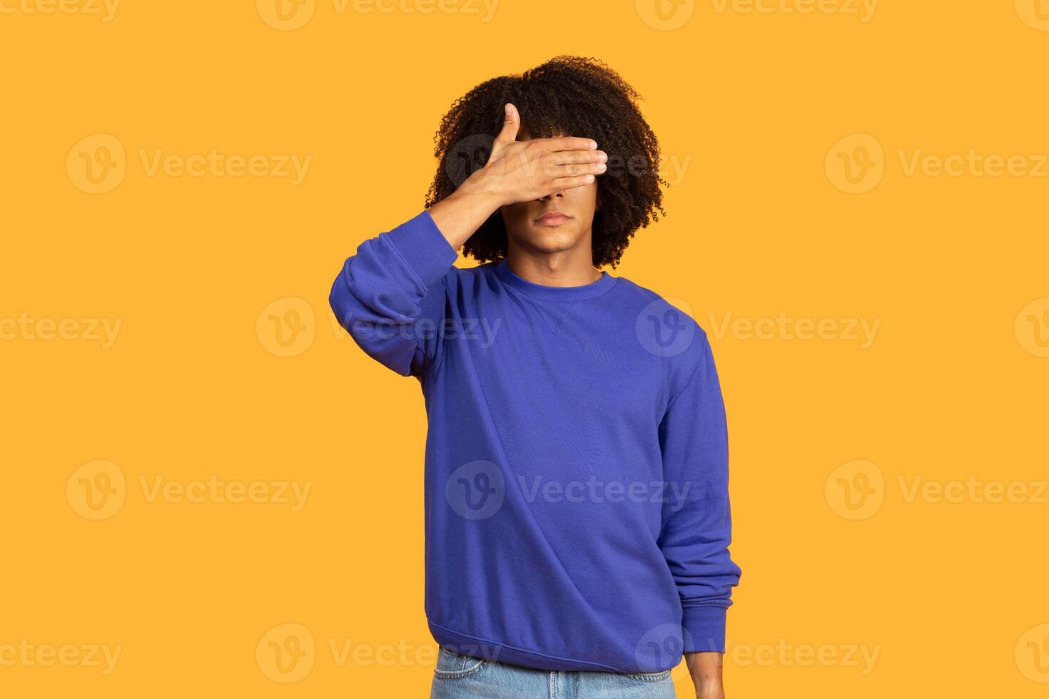 A man wearing a blue sweater is seen covering his eyes with his hands. He is standing against a neutral background, with a solemn expression on his face. photo