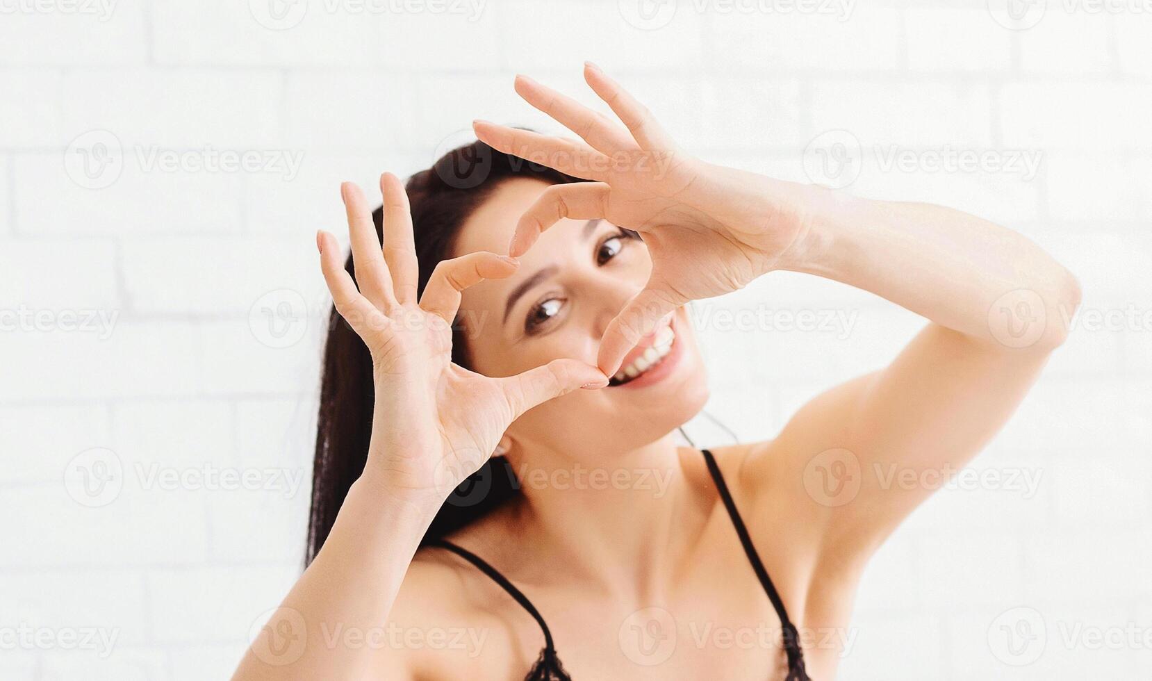 A cheerful woman wearing a black dress is making a heart shape with her hands while smiling. She is indoors, positioned against a bright, light-colored wall, and seems to be enjoying the moment. photo