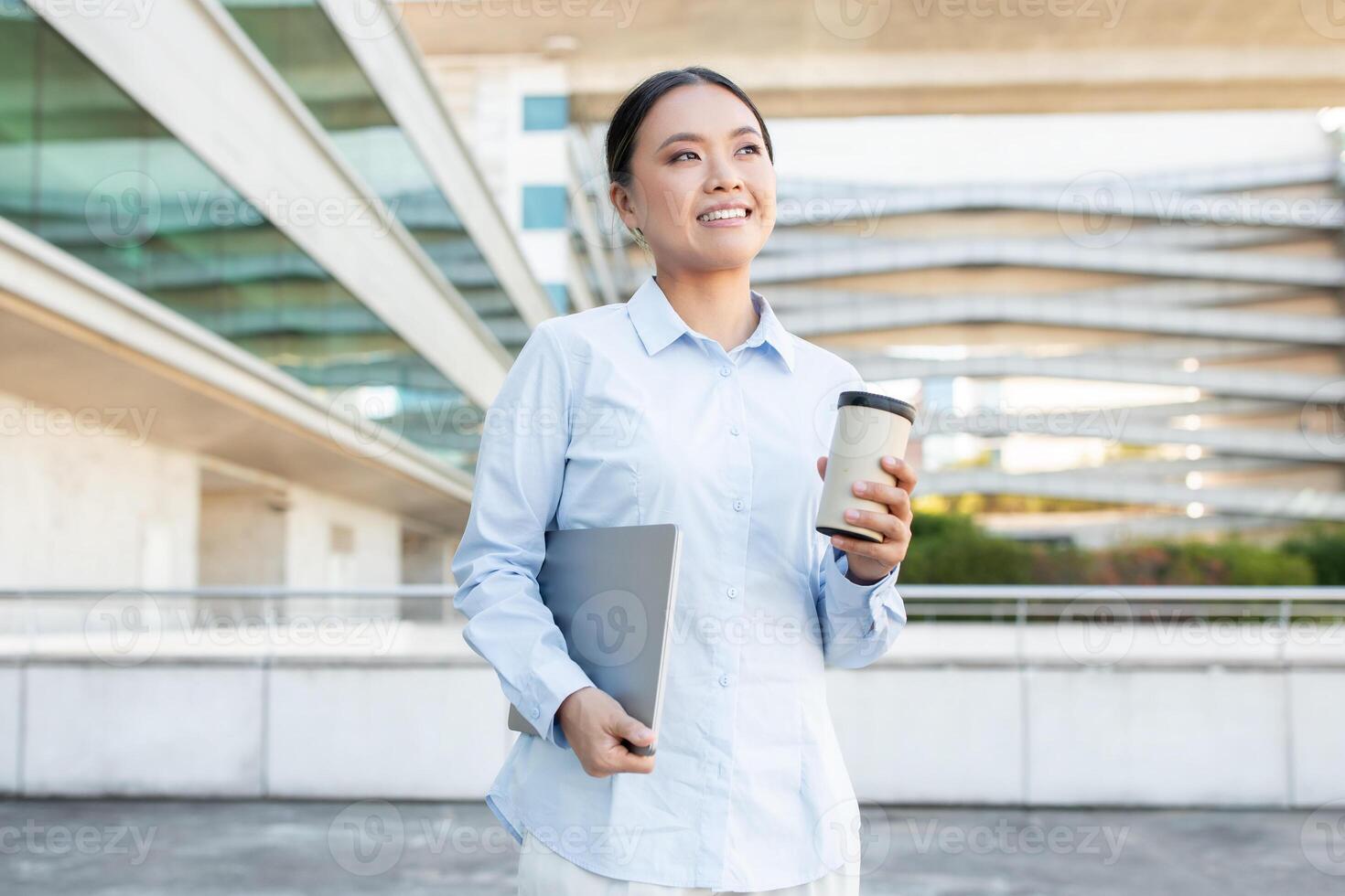 A young Asian woman stands outdoors in a sleek urban area, smiling brightly as she holds a coffee cup in one hand and a notebook in the other. photo