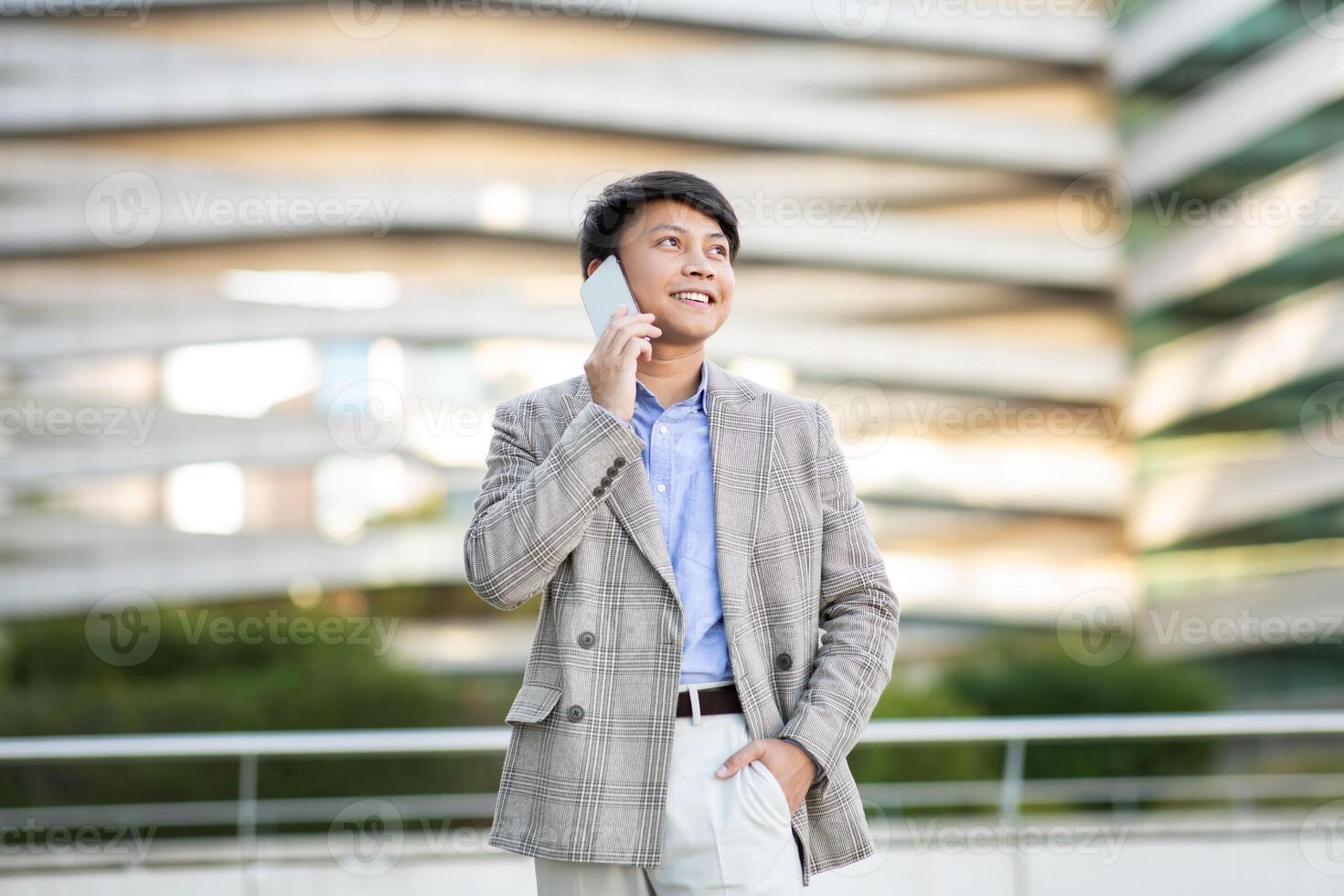 A young Asian man dressed in a grey plaid blazer and white pants is standing outside a modern building. He is smiling while talking on a white smartphone. photo