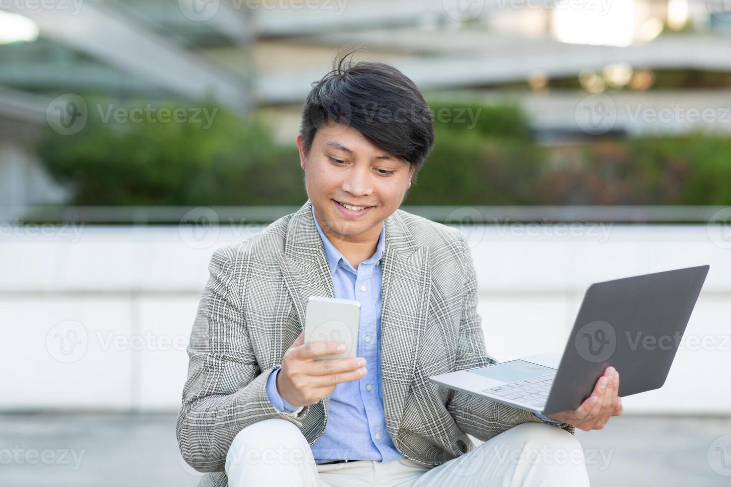 Asian man in a gray blazer and white pants sits on an outdoor ledge, smiling as he uses his phone. He is also holding a laptop on his lap. Green plants can be seen in the background. photo