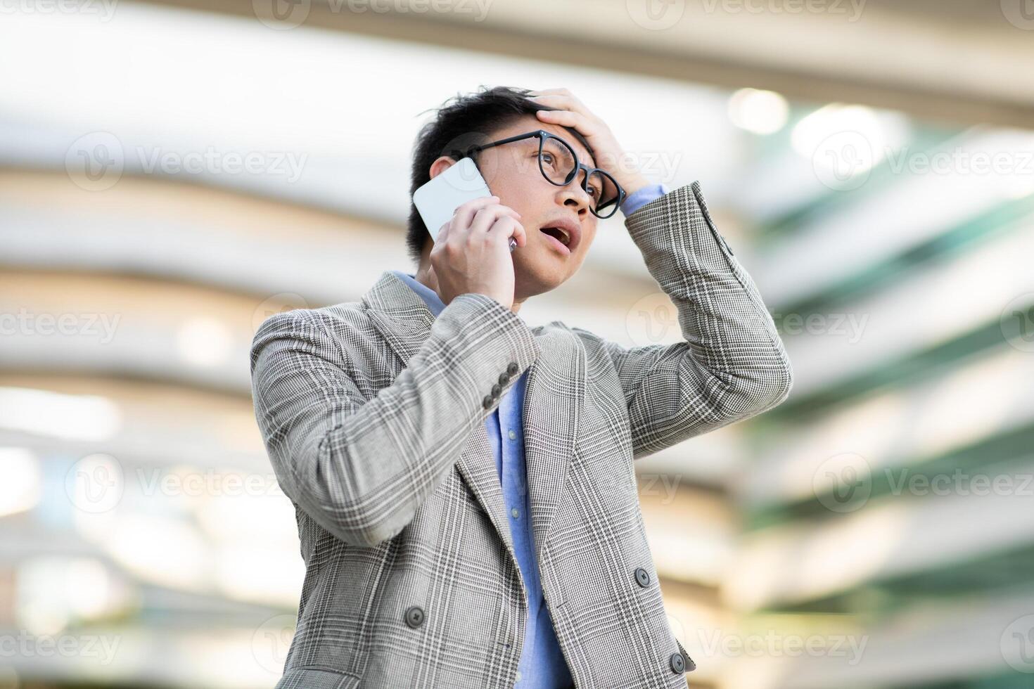 Asian man wearing glasses and a plaid blazer holds his head in his hand as he talks on a cell phone. He looks surprised and concerned. He is standing outside in front of a modern building. photo