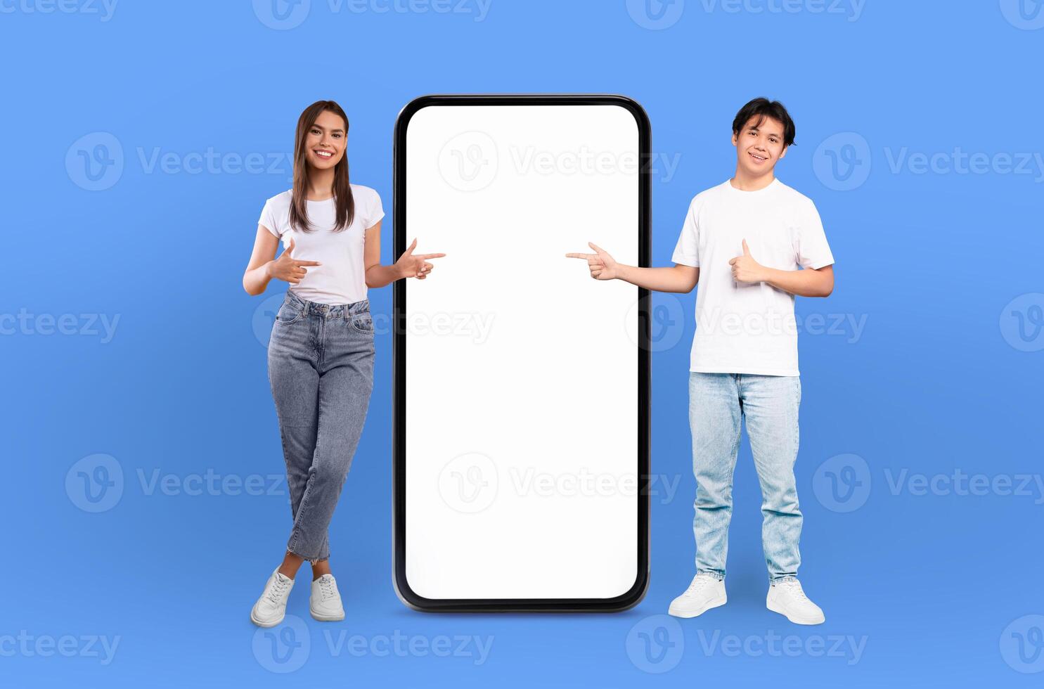 The scene features a young multiracial couple, both grinning, positioned next to a life-sized smartphone mockup on a seamless blue background. photo
