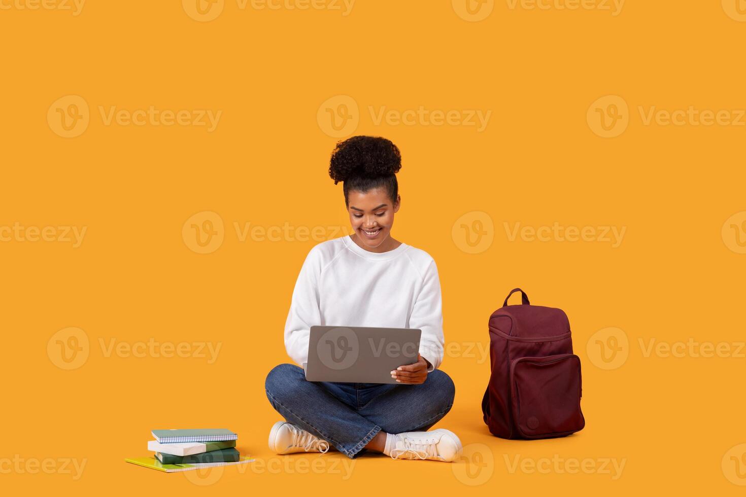 A young woman is sitting on a yellow background, using a laptop computer. A backpack is sitting on the floor next to her, and a stack of books is in front of her. photo