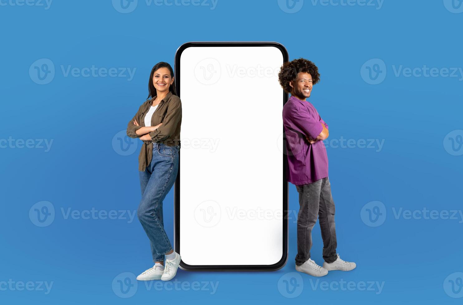 Smiling multiethnic couple leaning on an oversized, vertical smartphone with a blank white screen. They are both smiling and appear relaxed and happy, dressed in casual clothing photo