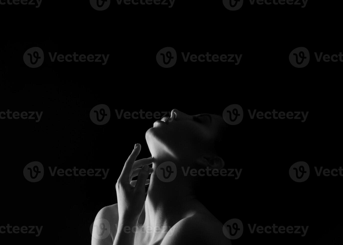 A black and white portrait of a woman with her head tilted upwards, touching her neck with her hand. The background is dark, creating a dramatic contrast, copy space above photo